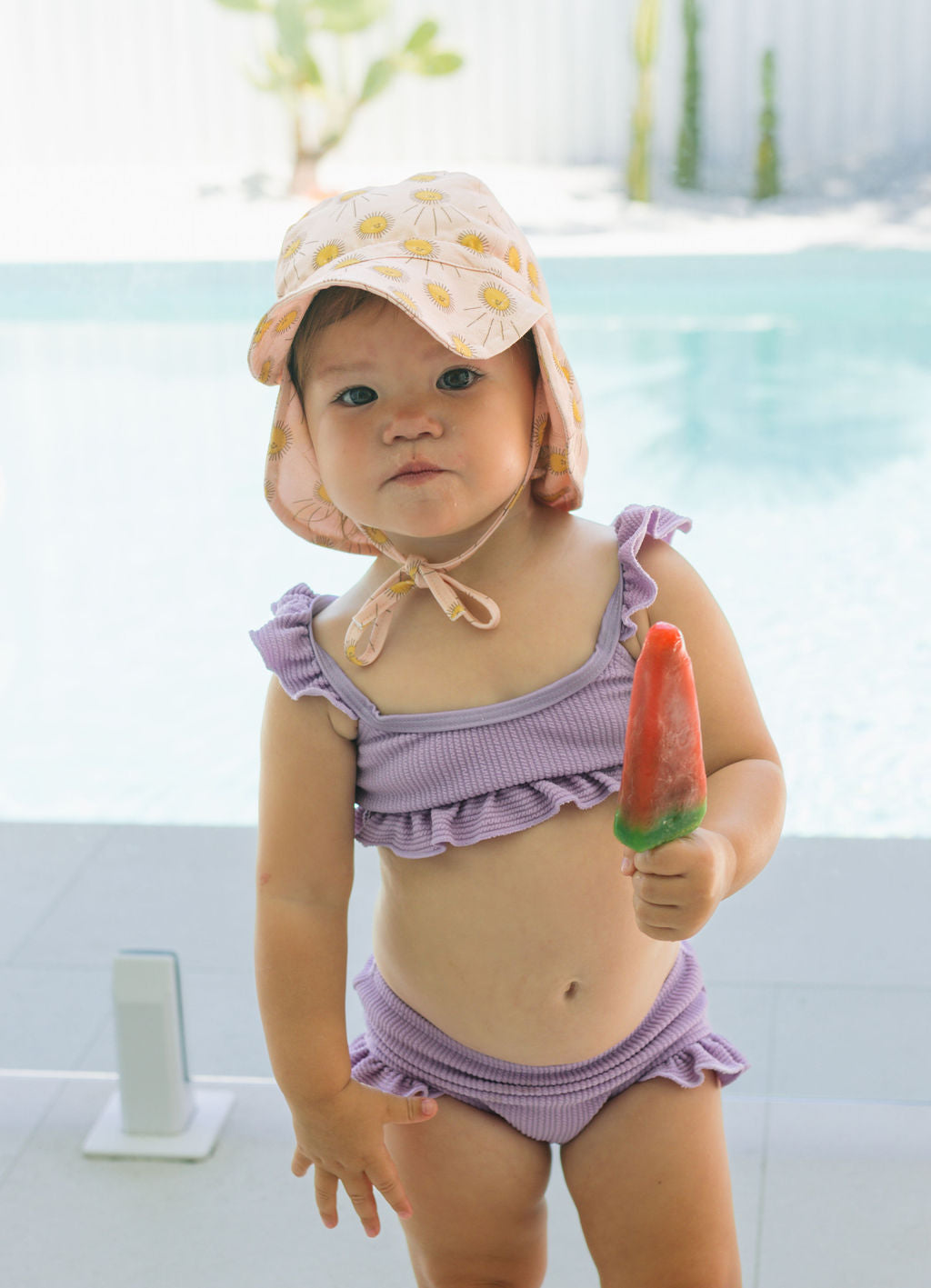 A toddler wears the ACORN KIDS Acorn Kids - Sunshine Flap Hat (UPF50+) and a lavender ruffled swimsuit by the pool, holding a red and green popsicle with a curious look on a bright sunny day with plants behind.