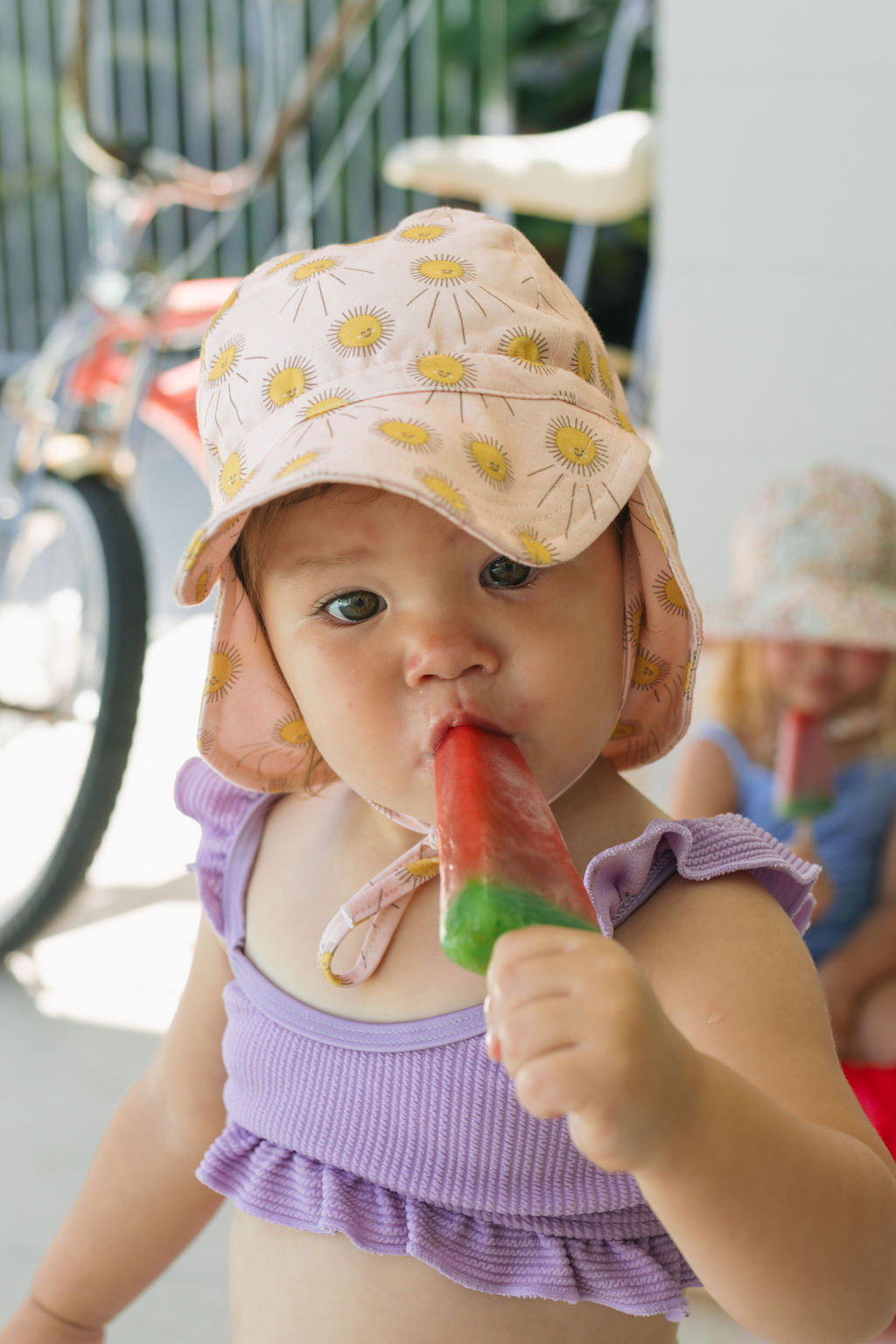 A toddler in an ACORN KIDS Acorn Kids - Sunshine Flap Hat (UPF50+, yellow flowers) and a purple cotton jersey top enjoys a red and green popsicle, while another child and a bicycle appear blurred in the background.