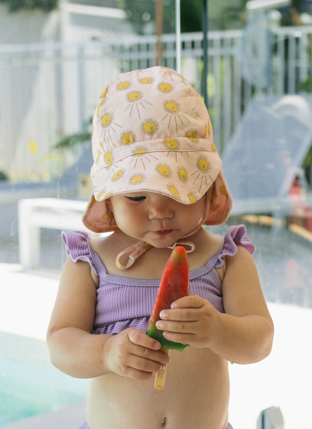 A toddler in a purple ruffled swimsuit and an ACORN KIDS Sunshine Flap Hat holds a red and green popsicle, gazing at it intently with a pool and lounge chair in the background.