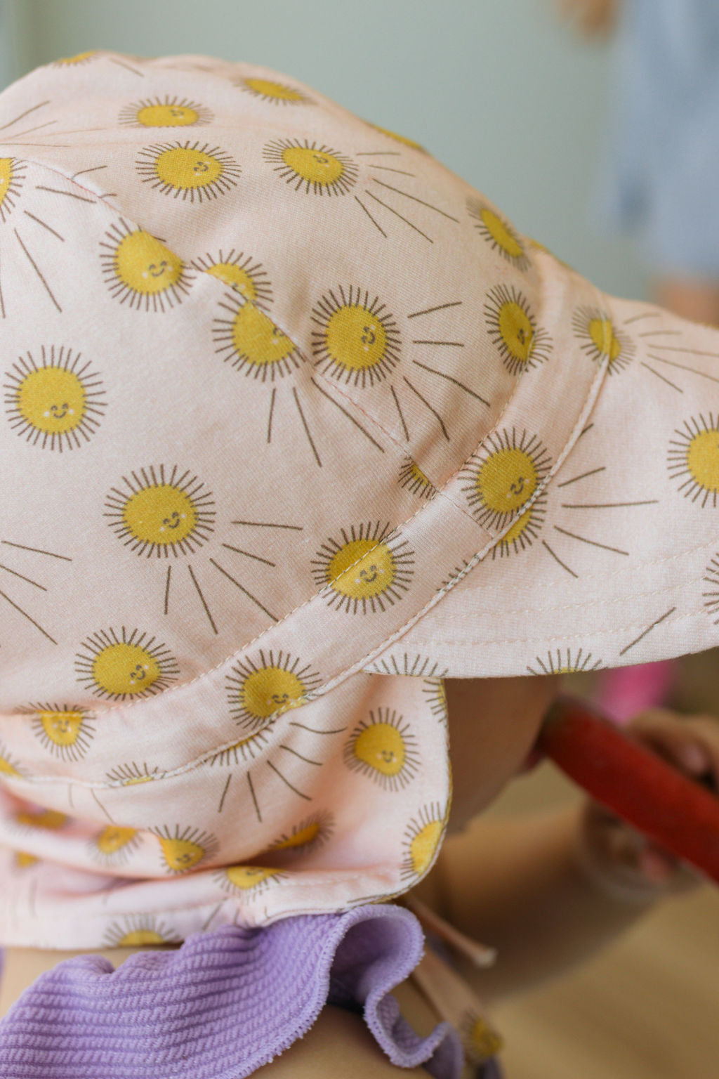 A child in purple and lilac wears the Acorn Kids - Sunshine Flap Hat by ACORN KIDS, a soft cotton jersey cap featuring yellow smiling suns. The UPF50+ hat shields from the sun while keeping the child’s face hidden as they hold a red object.