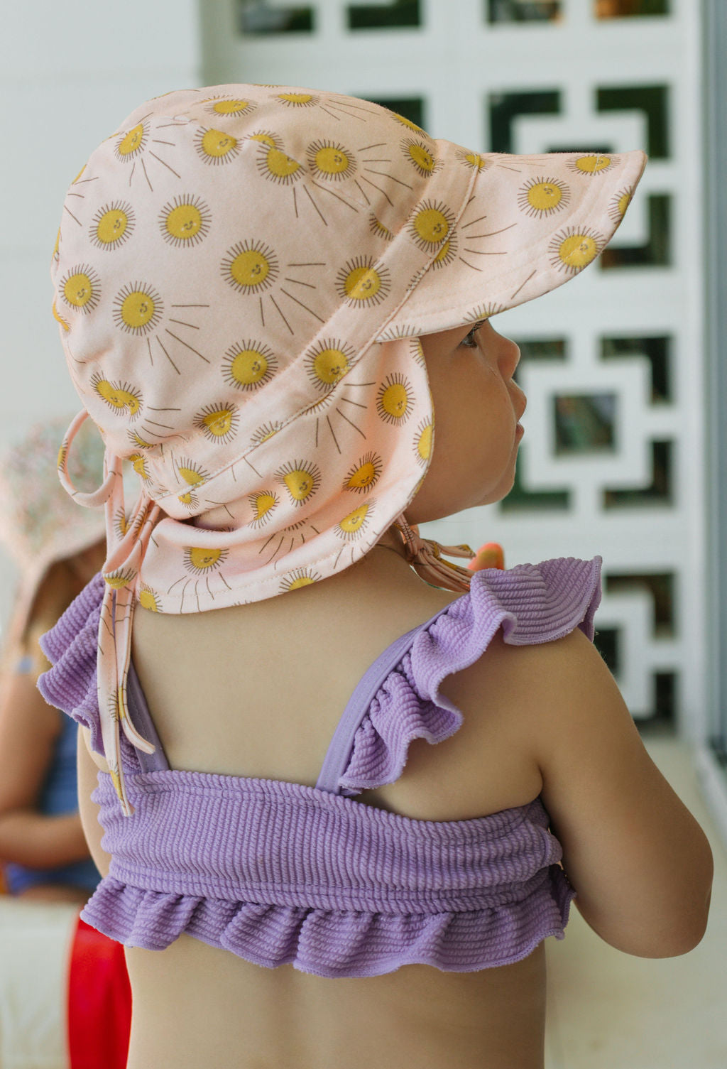 A young child in a purple ruffled top and an ACORN KIDS Sunshine Flap Hat with yellow daisies (UPF50+ sun protection) stands indoors, facing away from the camera. The background is softly blurred.