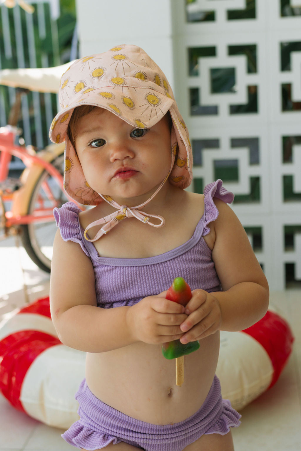 A toddler in a lavender ruffled swimsuit and the ACORN KIDS Sunshine Flap Hat holds a colorful popsicle indoors, with a red and white pool float and a bicycle in the background.