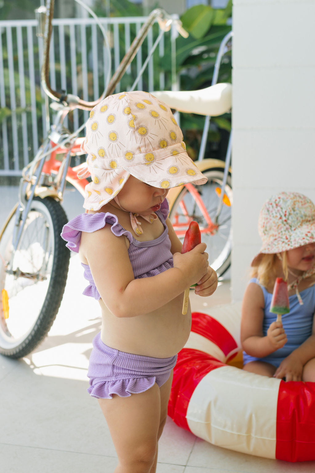Two kids in ACORN KIDS Acorn Kids - Sunshine Flap Hat and UPF50+ swimsuits enjoy popsicles indoors—one in a purple ruffled suit stands, while the other sits in a pool ring. A bicycle and green plants are behind them.