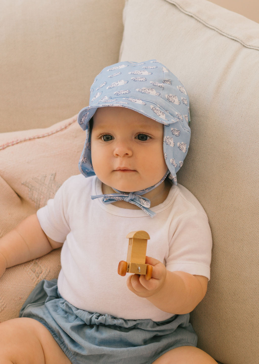 A baby in an ACORN KIDS Acorn Kids - Clouds Flap Hat, white shirt, and blue shorts sits on a light couch, holding a small wooden toy truck.