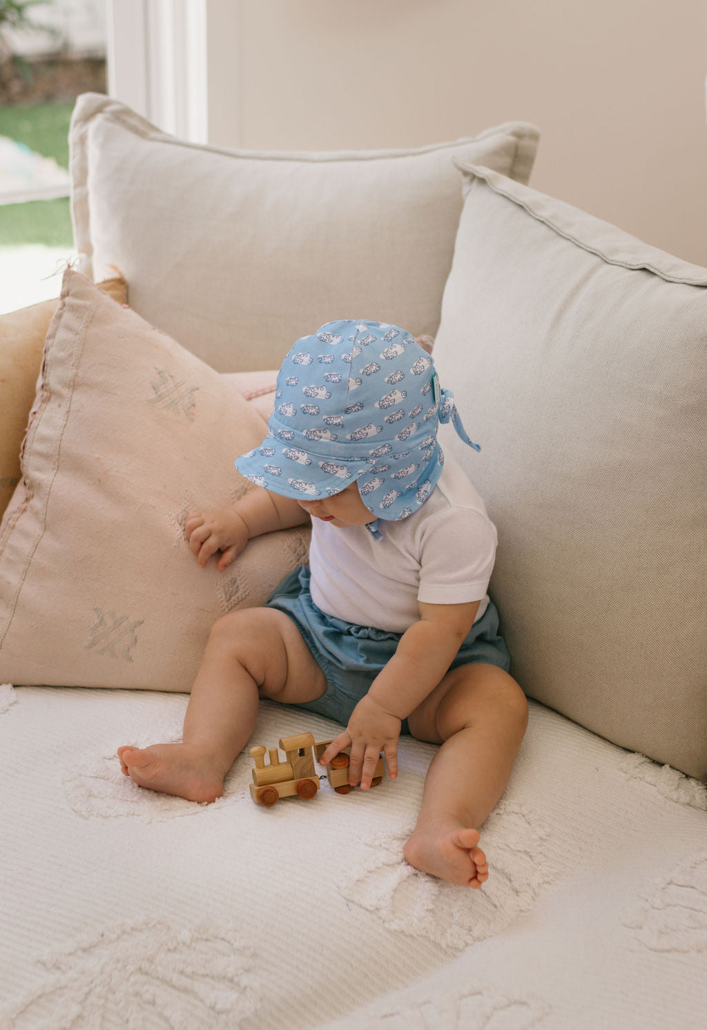 A baby wearing the ACORN KIDS Acorn Kids - Clouds Flap Hat with UPF50+ sun protection, a white shirt, and blue shorts sits on a beige couch, playing with a wooden toy truck as sunlight streams in through the window.