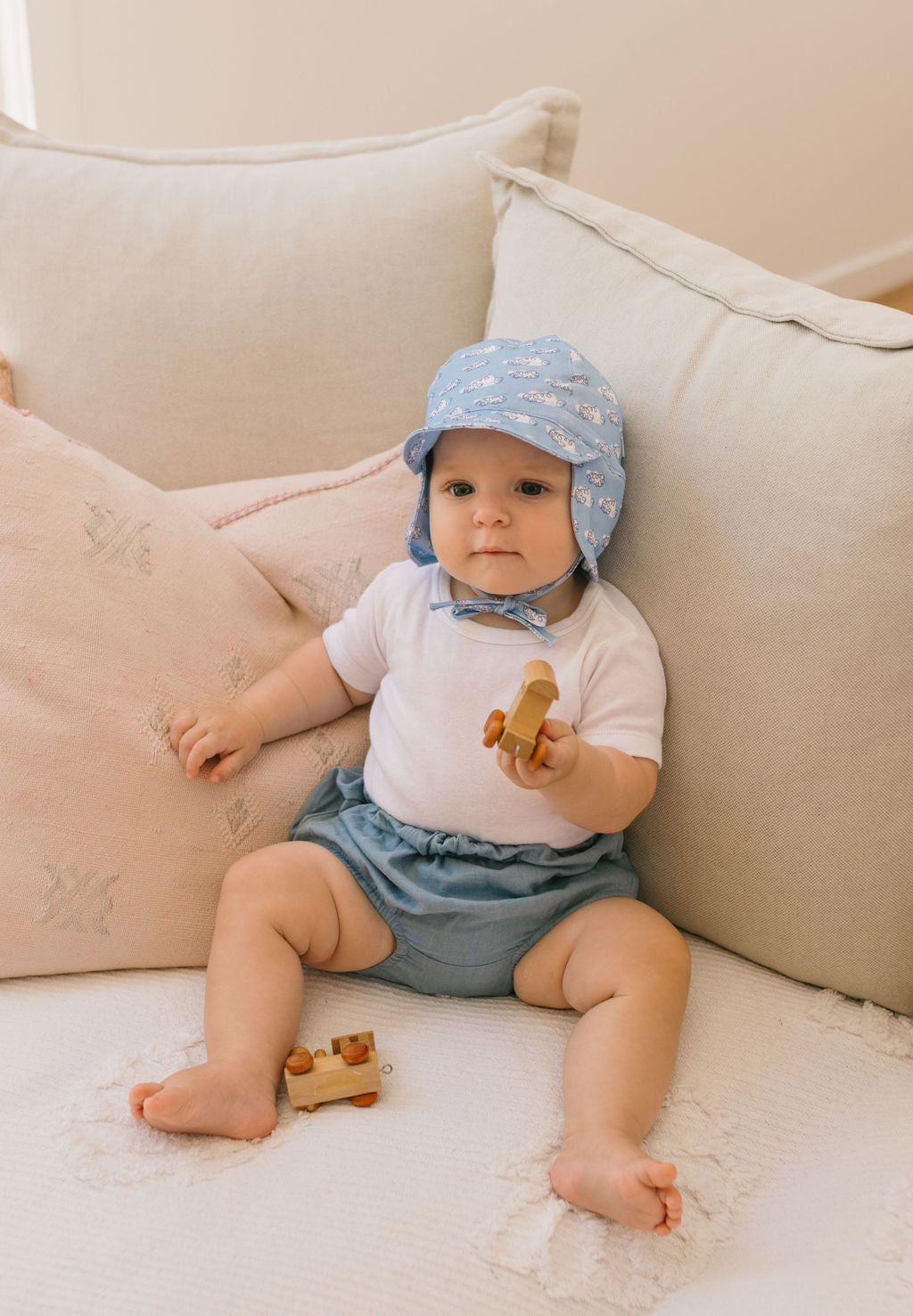 A baby in an ACORN KIDS Acorn Kids - Clouds Flap Hat (light blue, UPF50+), white shirt, and blue shorts sits on a beige couch, holding a wooden toy and surrounded by soft pillows.