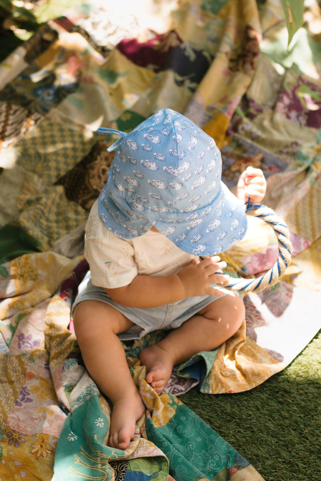 A baby in the ACORN KIDS Acorn Kids - Clouds Flap Hat with UPF50+ sun protection sits outdoors on a colorful quilt, looking down and holding a striped toy as sunlight filters through leaves.