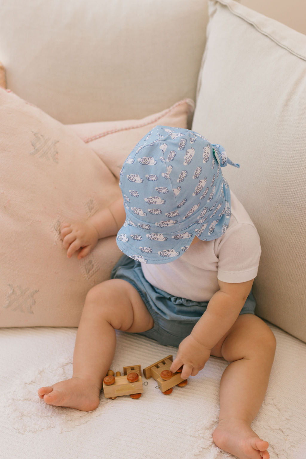 A baby in an ACORN KIDS Acorn Kids - Clouds Flap Hat, white shirt, and blue shorts sits on a light couch, holding a wooden toy truck with one hand on a pale pink pillow.