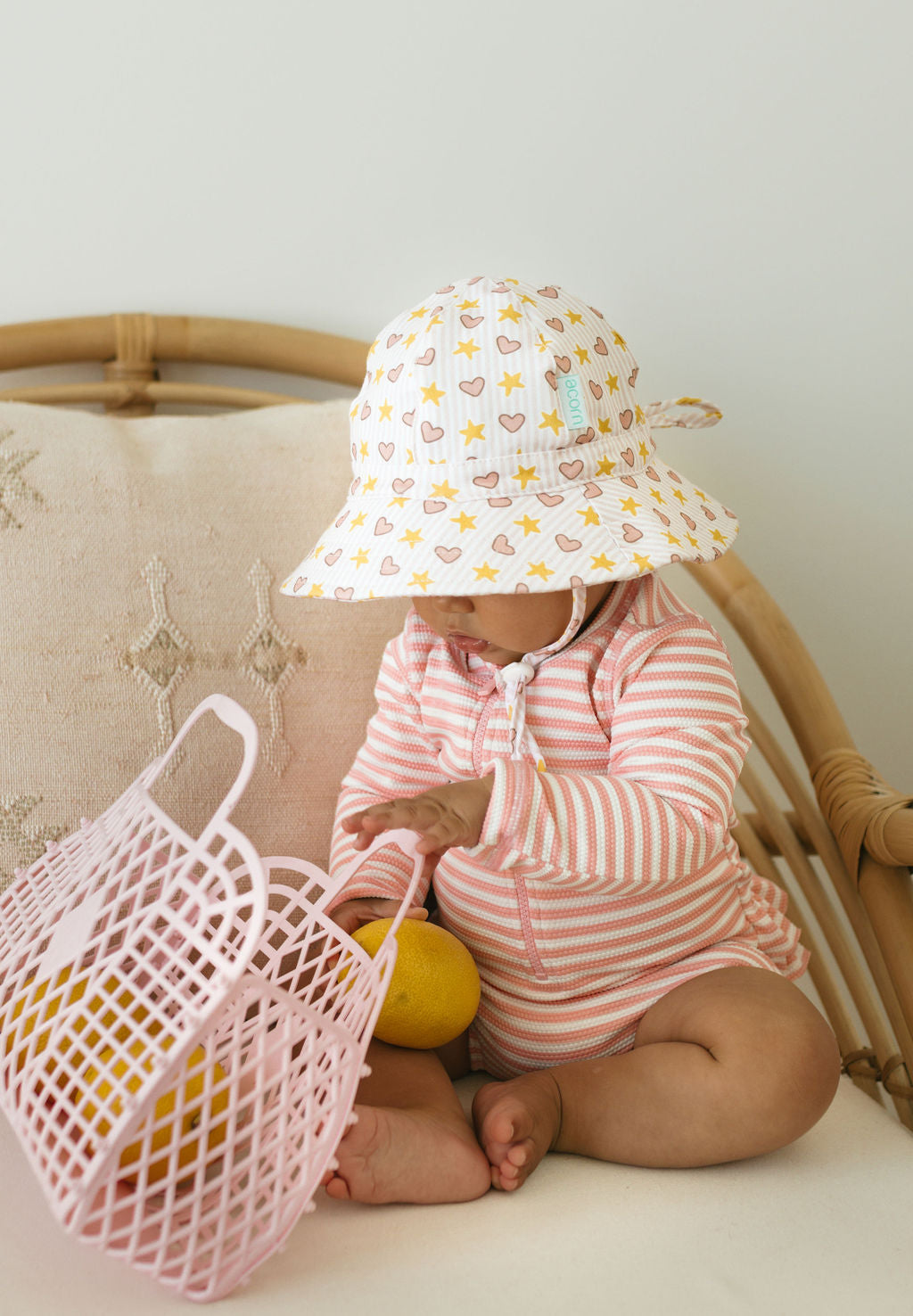 A baby in a striped cotton jersey outfit and an ACORN KIDS Acorn Kids - Love and Stars Stretch Cotton Wide Brim Sunhat sits on a cushioned chair, placing yellow fruit in a pink plastic basket.