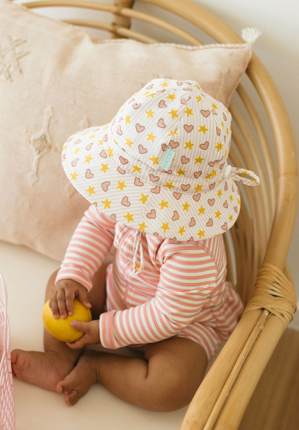 A baby in a striped cotton romper and the ACORN KIDS Love and Stars Stretch Cotton Wide Brim Sunhat sits on a wicker chair, holding a lemon. A beige pillow embroidered with stars appears in the background.