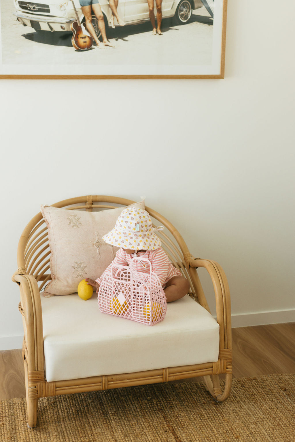 A young child in the Acorn Kids - Love and Stars Stretch Cotton Wide Brim Sunhat by ACORN KIDS sits on a wicker chair with a beige cushion, holding a pink bag. A lemon is on the seat, and a framed beach photo hangs above.
