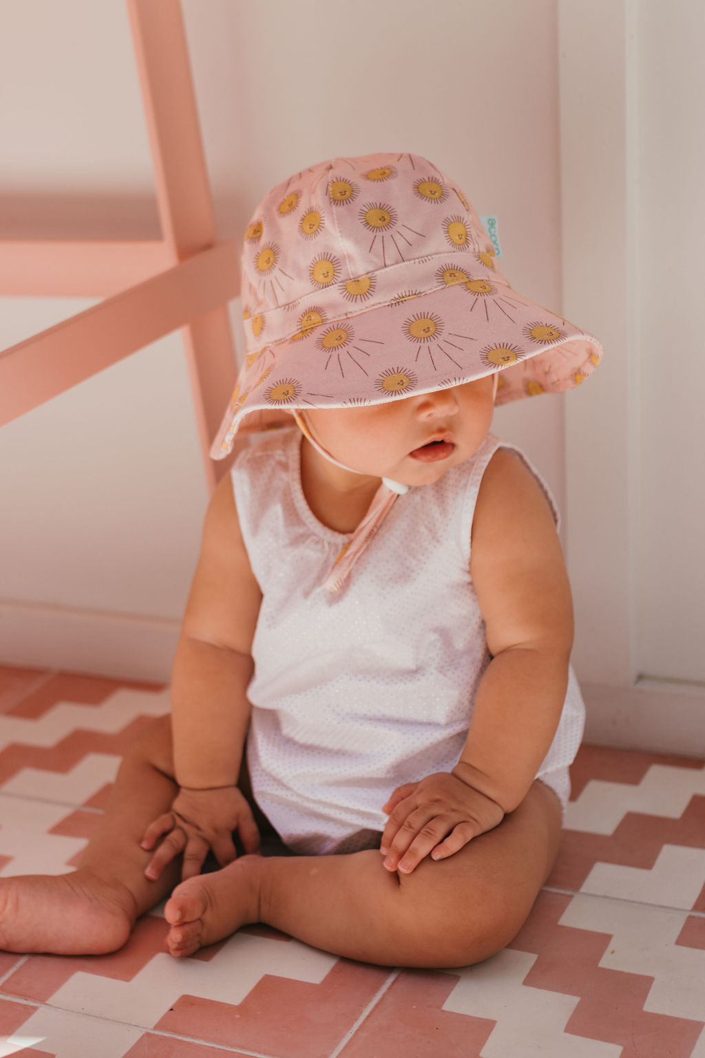 A baby in the ACORN KIDS Sunshine Stretch Cotton Wide Brim Sunhat sits on patterned pink and white floor tiles, leaning against a pink chair indoors, wearing a pale pink sleeveless outfit.