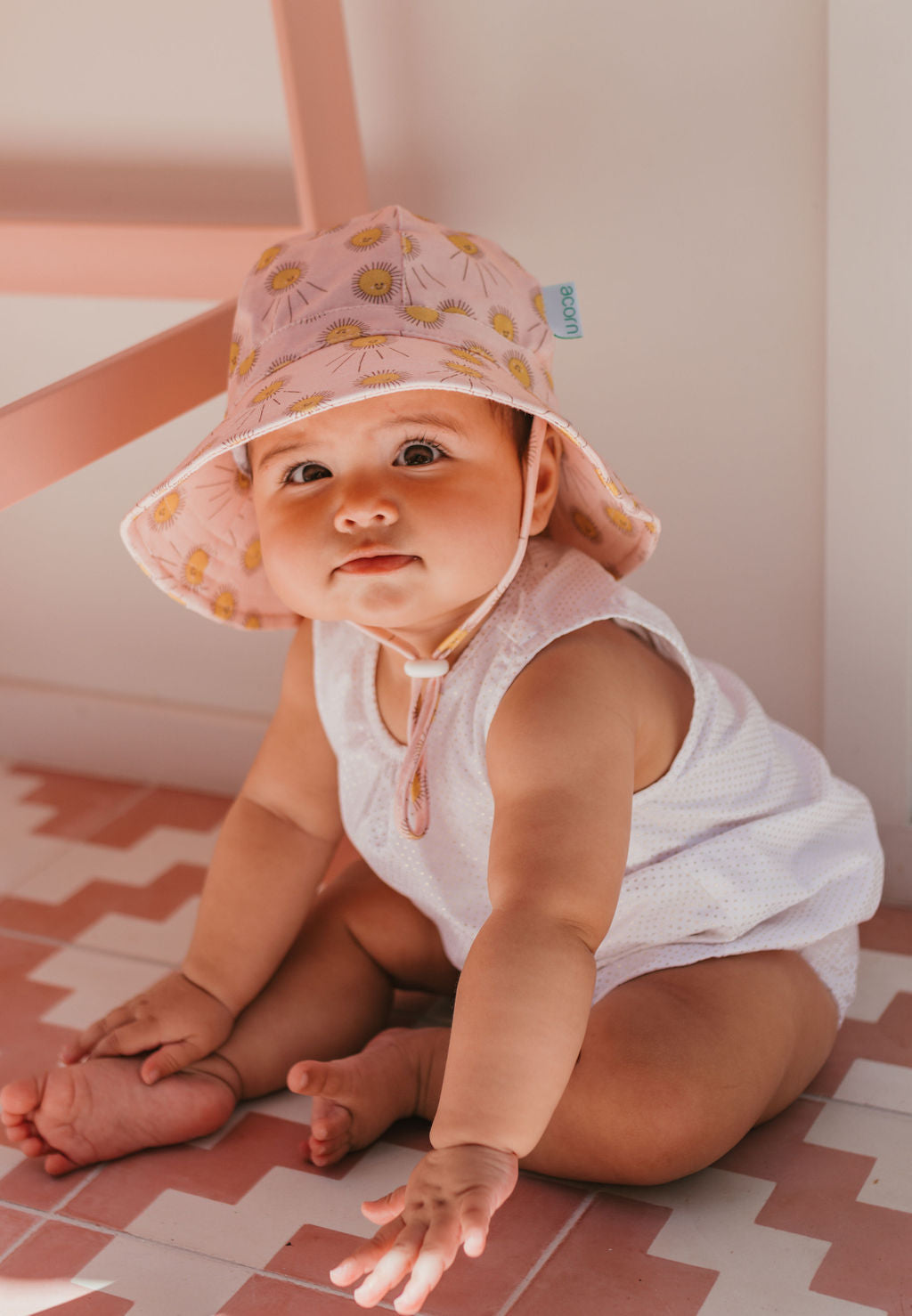 A baby in a white outfit wears the ACORN KIDS Sunshine Stretch Cotton Wide Brim Sunhat, featuring yellow flower prints, and sits on a tiled floor reaching forward with a curious look.
