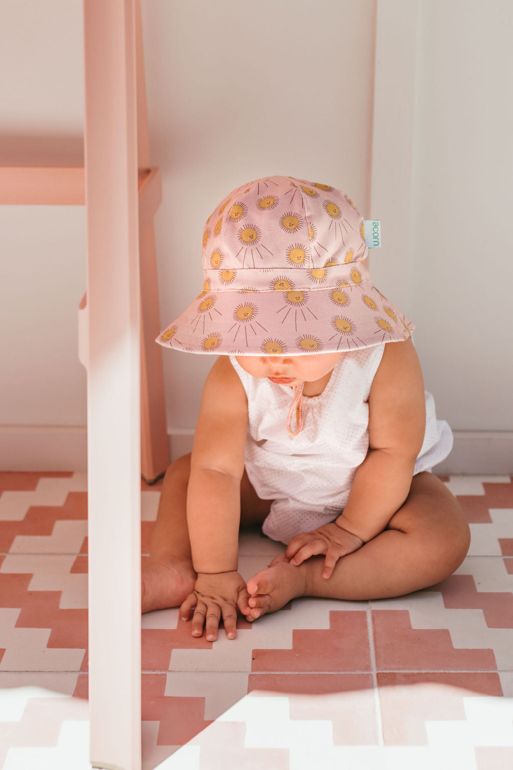 A baby in an ACORN KIDS Sunshine Stretch Cotton Wide Brim Sunhat with yellow flowers sits on a pink and white tiled floor, dressed in white and touching the floor while looking down.