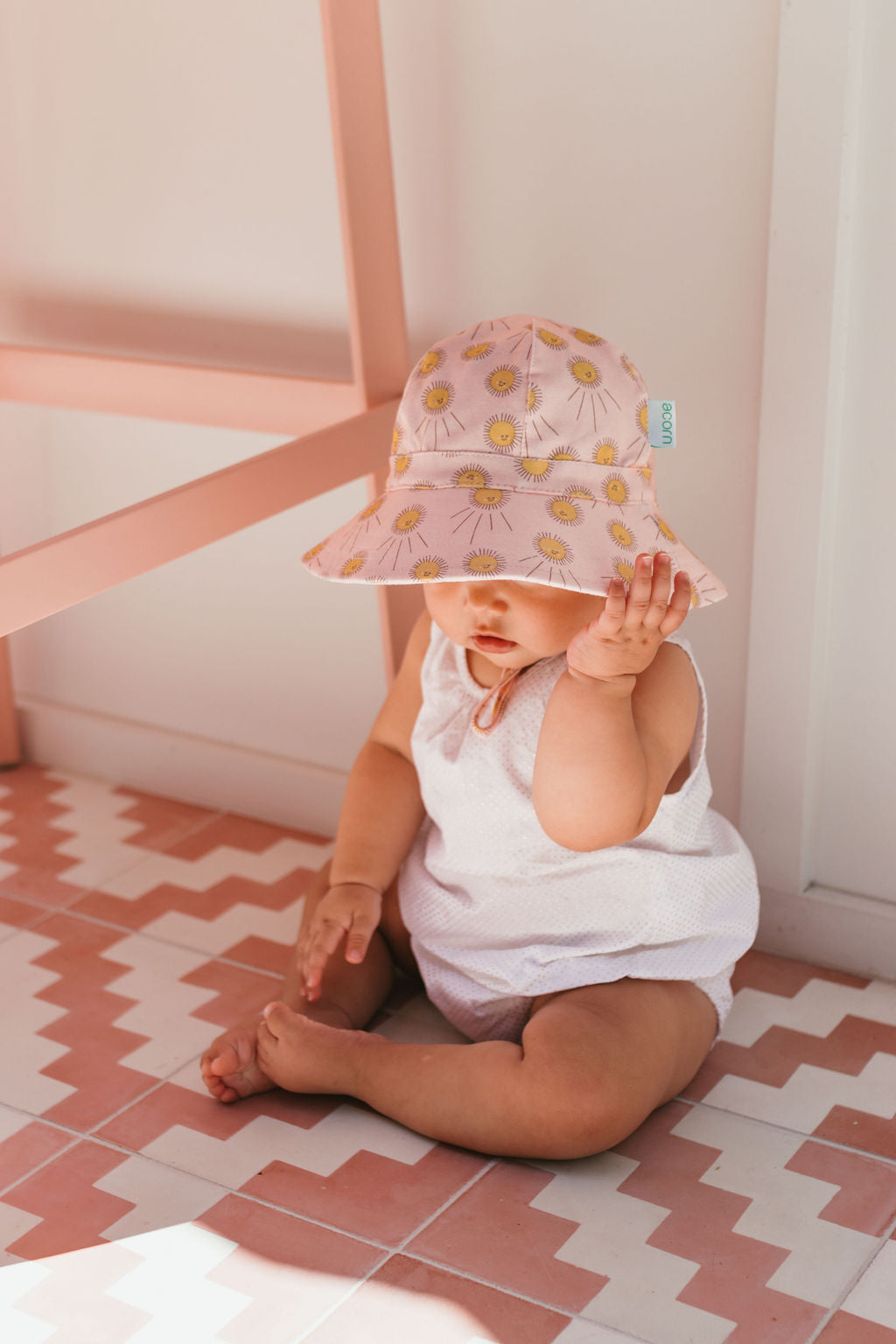 A baby wearing the ACORN KIDS Acorn Kids - Sunshine Stretch Cotton Wide Brim Sunhat with UPF50+ sits barefoot on a pink and white zigzag floor, dressed in white and raising one hand near their face.