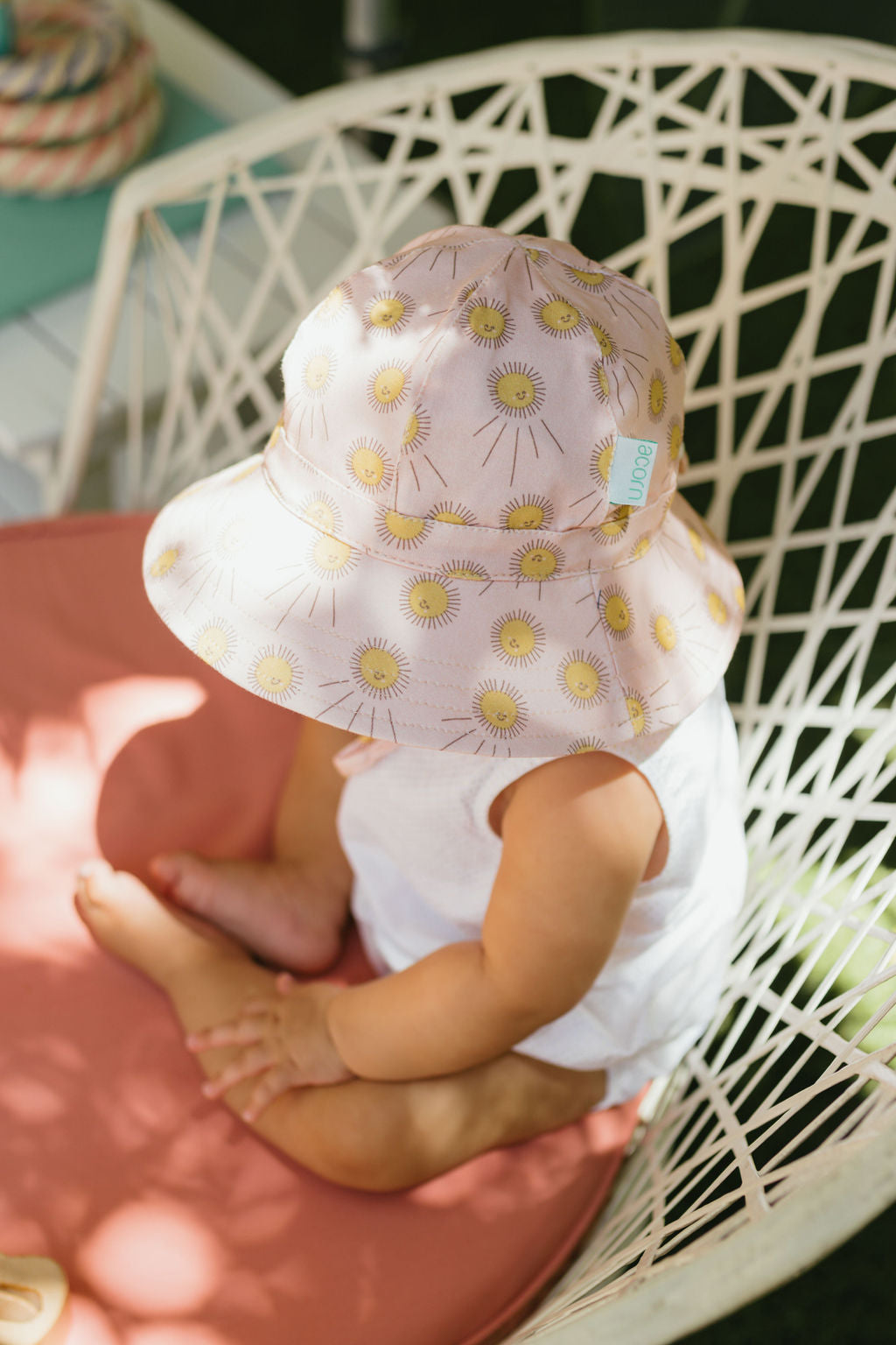 A baby wearing a white outfit and the ACORN KIDS Sunshine Stretch Cotton Wide Brim Sunhat with UPF50+ sits on a peach cushion in a patterned white chair, viewed from above.
