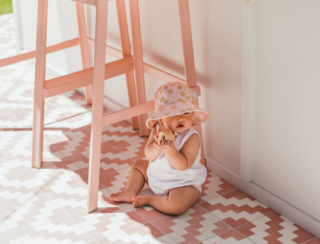 A baby in an ACORN KIDS Acorn Kids - Sunshine Stretch Cotton Wide Brim Sunhat and white outfit sits on a pink and white patterned tile floor beside a pink chair, sunlight streaming in.