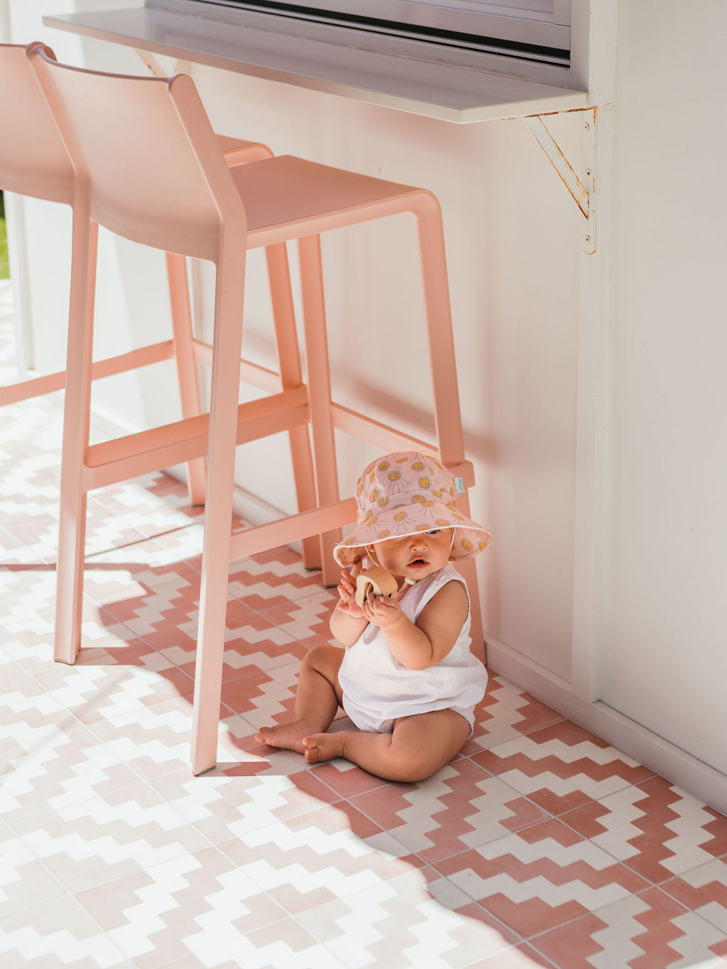A baby in the ACORN KIDS Acorn Kids - Sunshine Stretch Cotton Wide Brim Sunhat with UPF50+ protection and a white outfit smiles and plays on a pink-and-white tile floor beside a pink bar stool in a bright, sunny room.