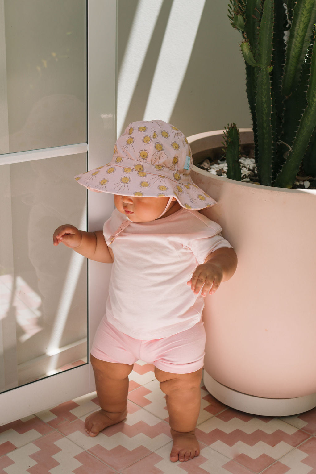 A baby in an ACORN KIDS Acorn Kids - Sunshine Stretch Cotton Wide Brim Sunhat stands indoors by a large potted cactus, sunlight streaming through a glass door.