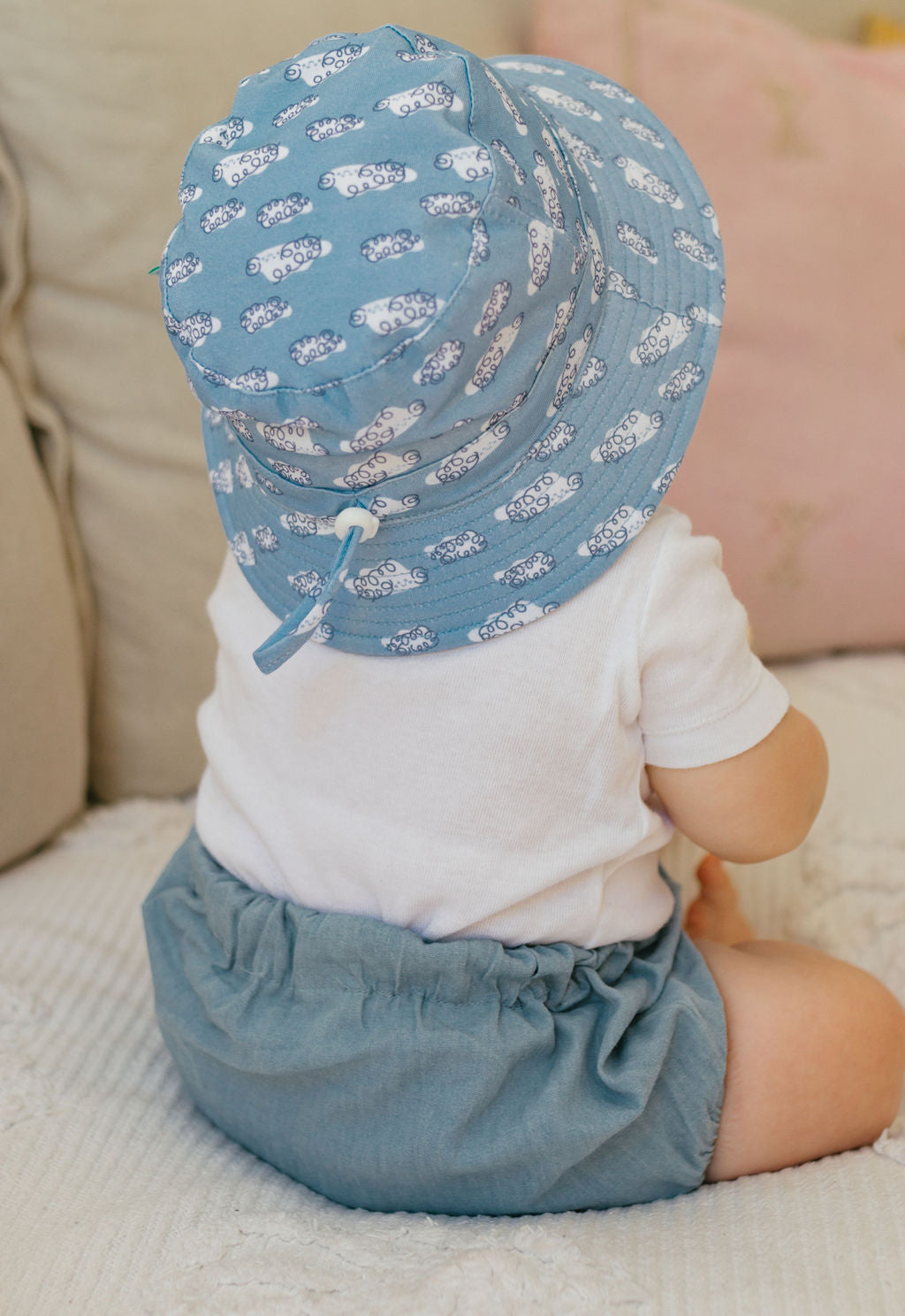 A baby sits on a light blanket, wearing a white shirt, blue shorts, and the ACORN KIDS Acorn Kids - Clouds Stretch Cotton Wide Brim Sunhat with UPF50+ protection. A beige and pink pillow is in the background.