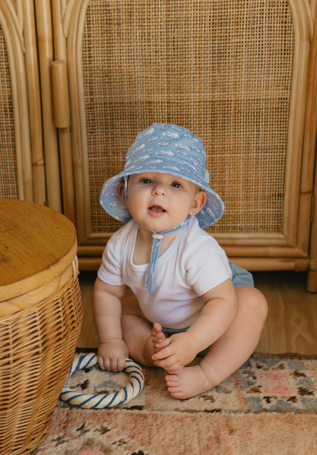 A baby in an ACORN KIDS Acorn Kids - Clouds Stretch Cotton Wide Brim Sunhat with UPF50+ and a white onesie sits on a patterned rug by a wicker basket, holding a rope toy and looking at the camera.
