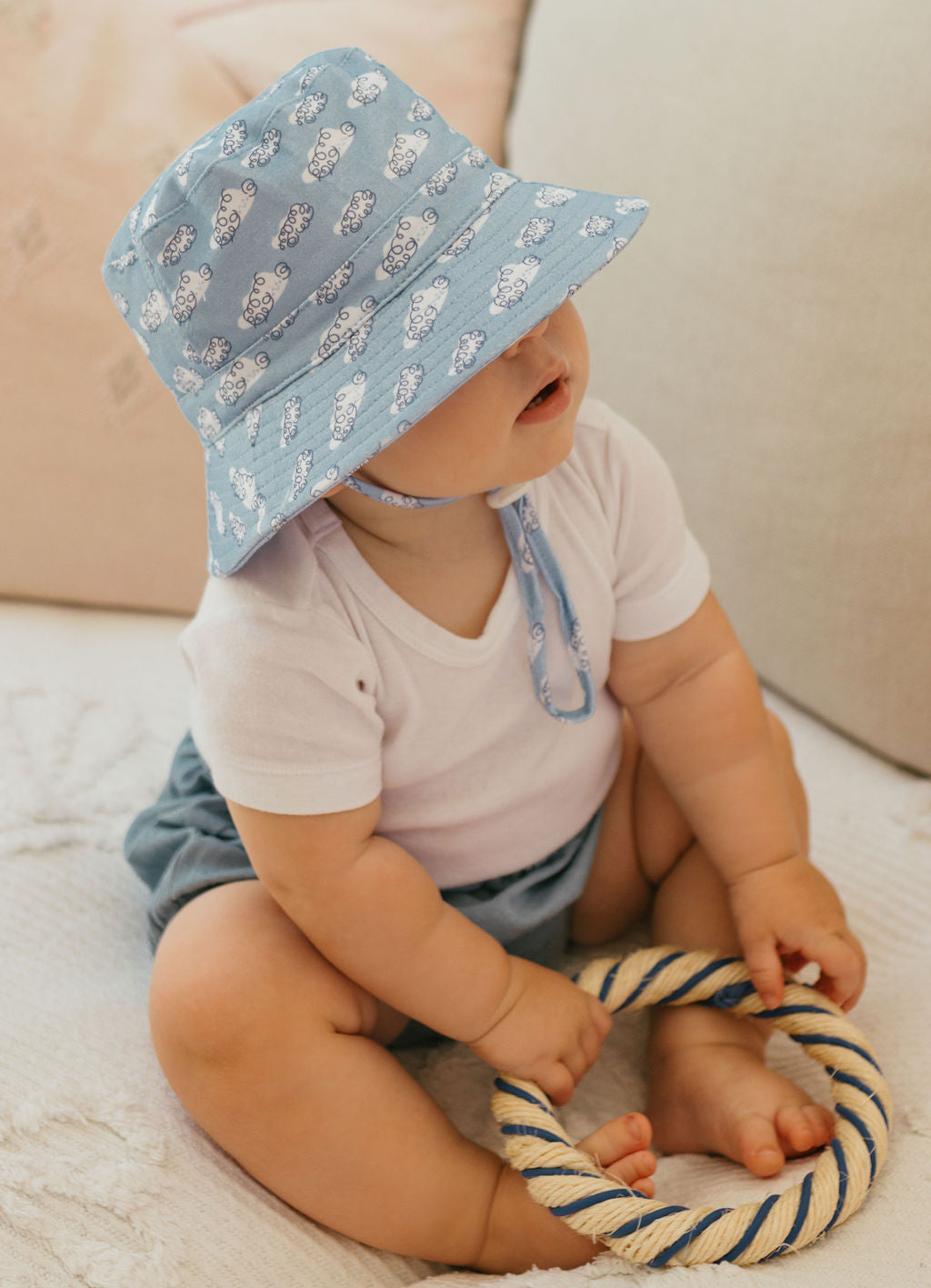 A baby in the ACORN KIDS Acorn Kids - Clouds Stretch Cotton Wide Brim Sunhat sits on a light blanket, holding a round rope toy. Dressed in a white shirt and blue shorts, the baby enjoys UPF50+ sun protection.