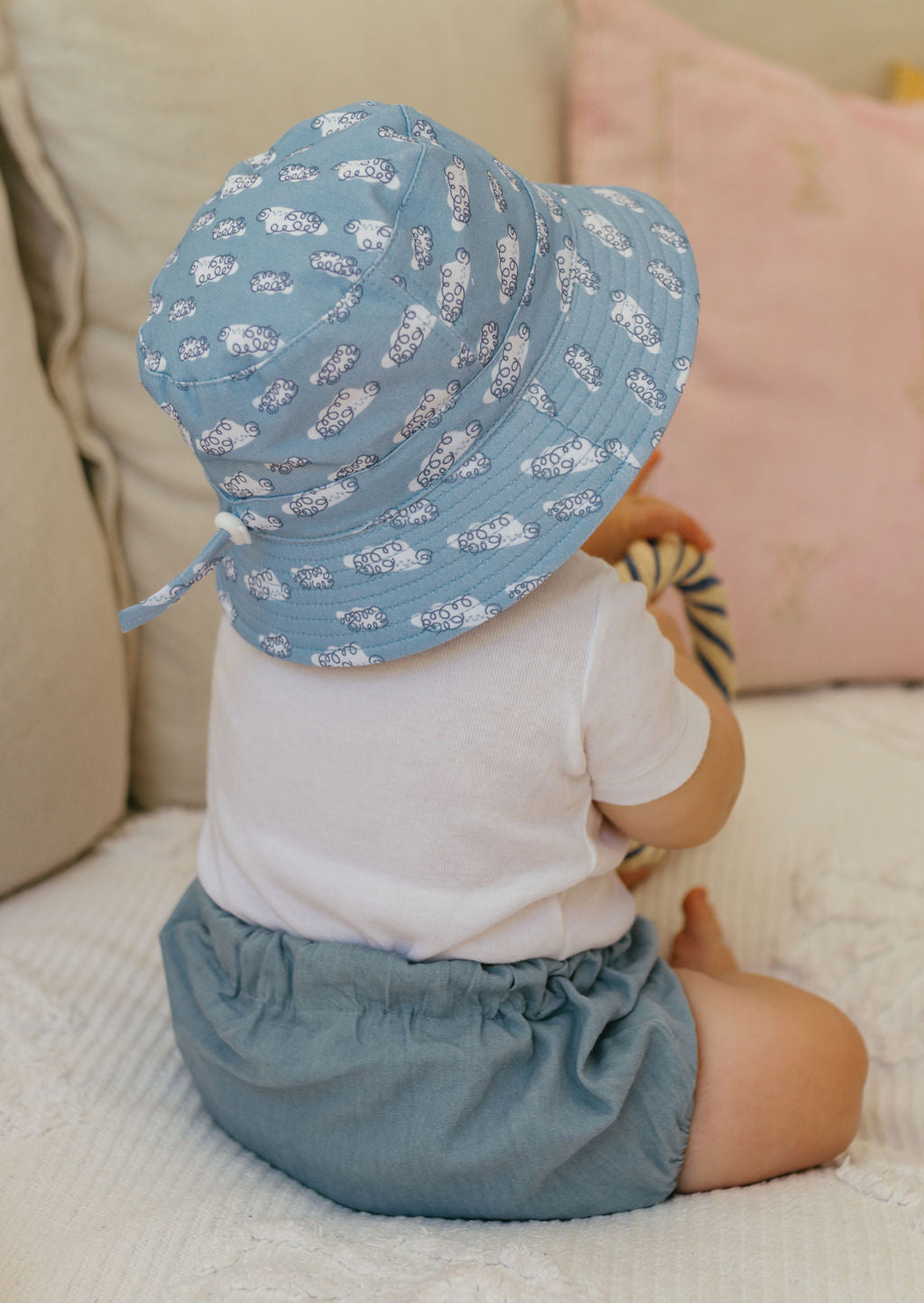 A baby wearing the Acorn Kids - Clouds Stretch Cotton Wide Brim Sunhat by ACORN KIDS, a white shirt, and blue shorts sits on a bed facing away, holding a toy. Cream and pink cushions are in the background.