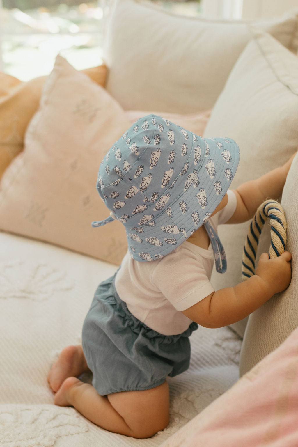 A baby wearing the ACORN KIDS Acorn Kids - Clouds Stretch Cotton Wide Brim Sunhat kneels on a cream couch, holding a rope-detailed cushion, dressed in a white shirt and blue shorts.
