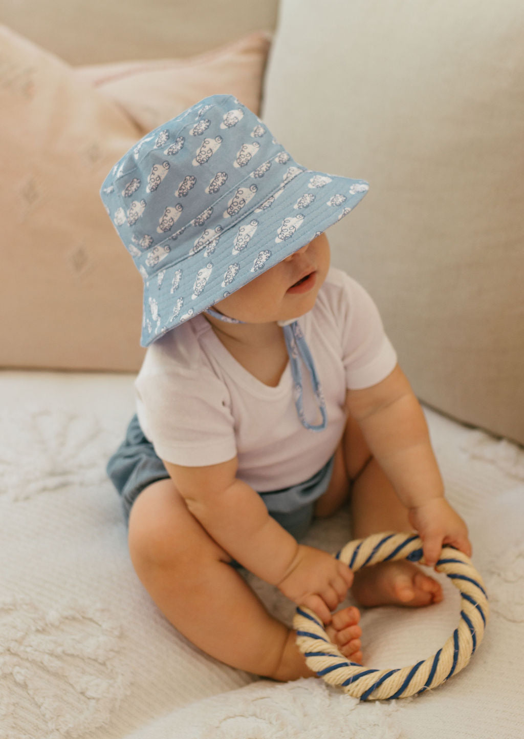 A baby in an ACORN KIDS Clouds Stretch Cotton Wide Brim Sunhat and a white shirt sits on a sofa, holding a rope ring toy.
