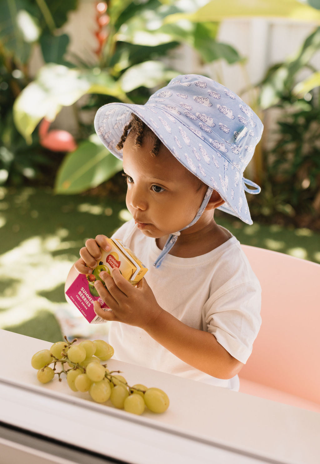 A young child in an ACORN KIDS Acorn Kids - Clouds Stretch Cotton Wide Brim Sunhat (light blue, UPF50+) and white shirt sits outdoors sipping juice, with green grapes in front and lush plants in the background.
