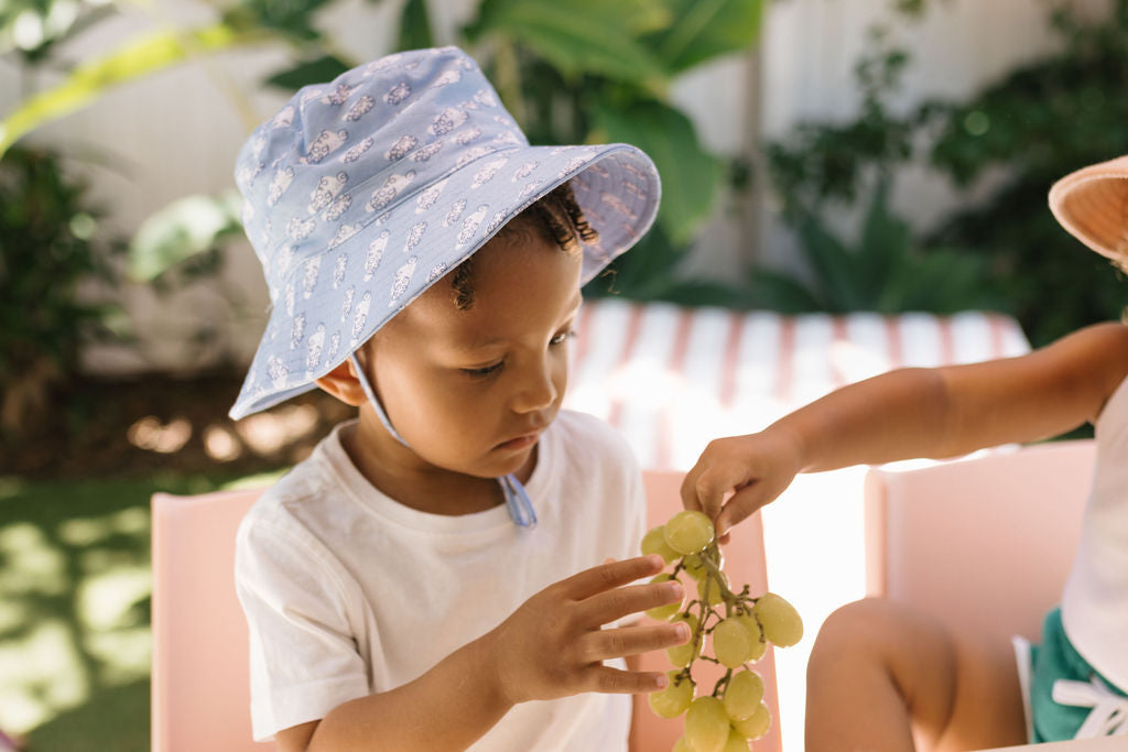 A young child in the ACORN KIDS Acorn Kids - Clouds Stretch Cotton Wide Brim Sunhat with UPF50+ and a white shirt holds green grapes, while another child's hand reaches in. They are outdoors with greenery in the background.