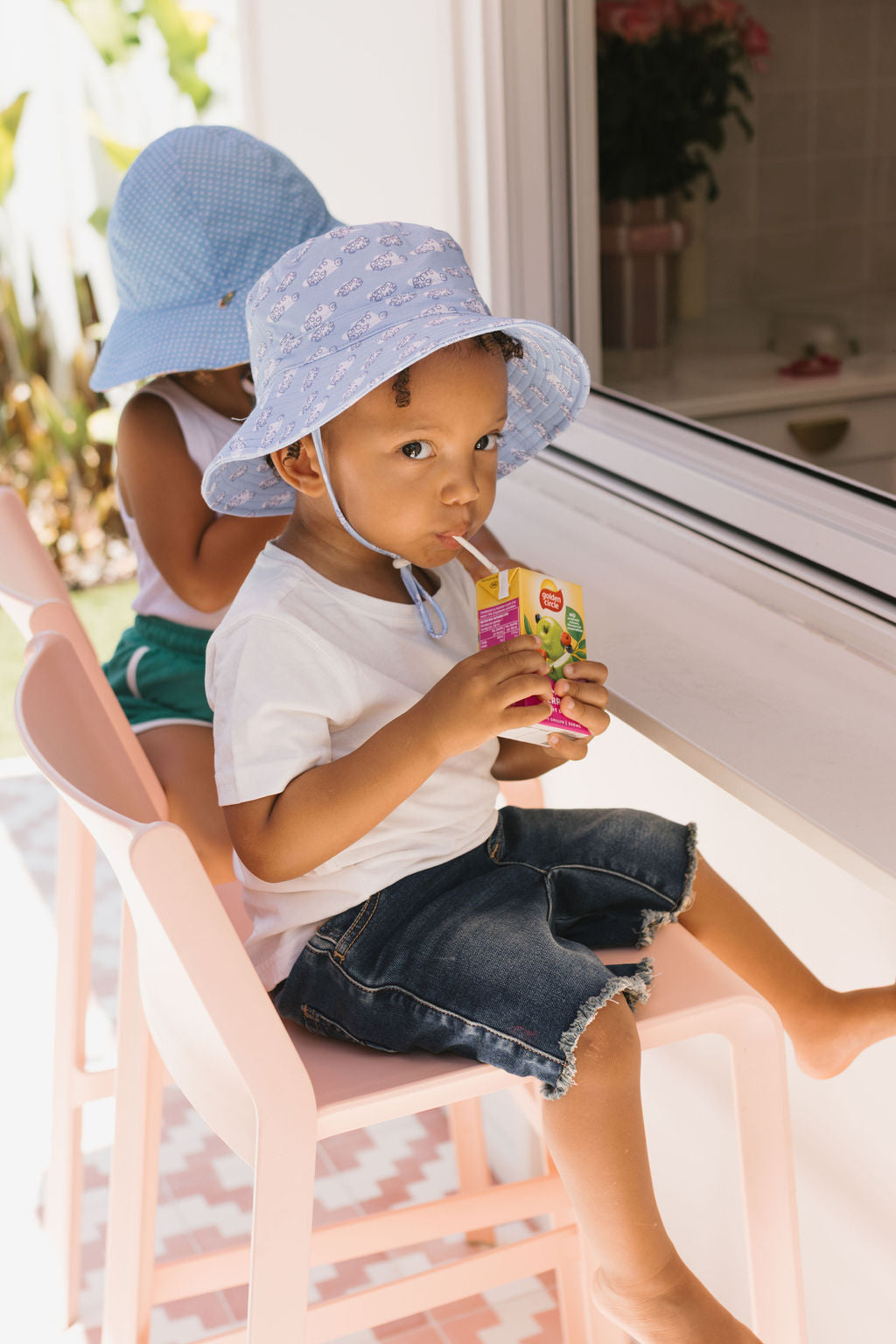 Two young children sit by a window. The child in front, in a wide brim blue sun hat and white shirt, drinks from a juice box. Behind, the other wears an ACORN KIDS Clouds Stretch Cotton Sunhat with UPF50+ protection, facing away.