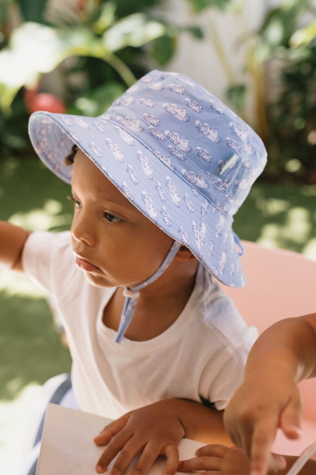 A young child in a white shirt wears the ACORN KIDS "Acorn Kids - Clouds Stretch Cotton Wide Brim Sunhat" with UPF50+ protection, sitting outdoors among green plants and sunlight as another hand points nearby.