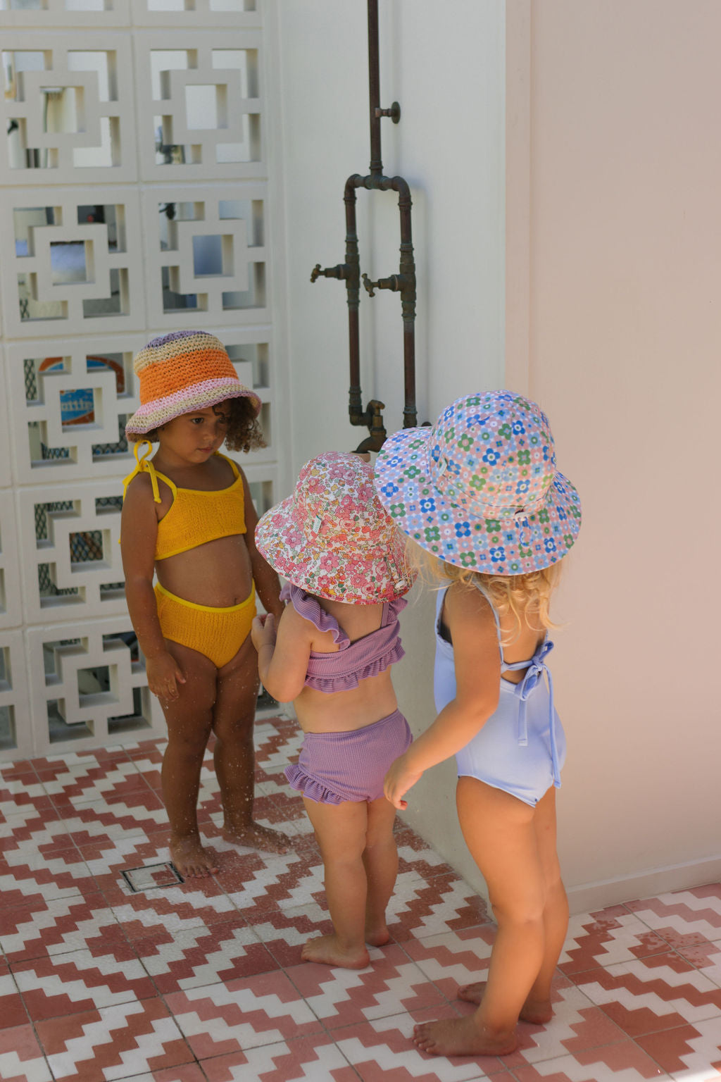 Three young children in swimsuits wear ACORN KIDS Acorn Kids - Tessa Broad Brim Bucket Hat (UPF50+) by an outdoor shower on a tiled floor, with a decorative white wall in the background.