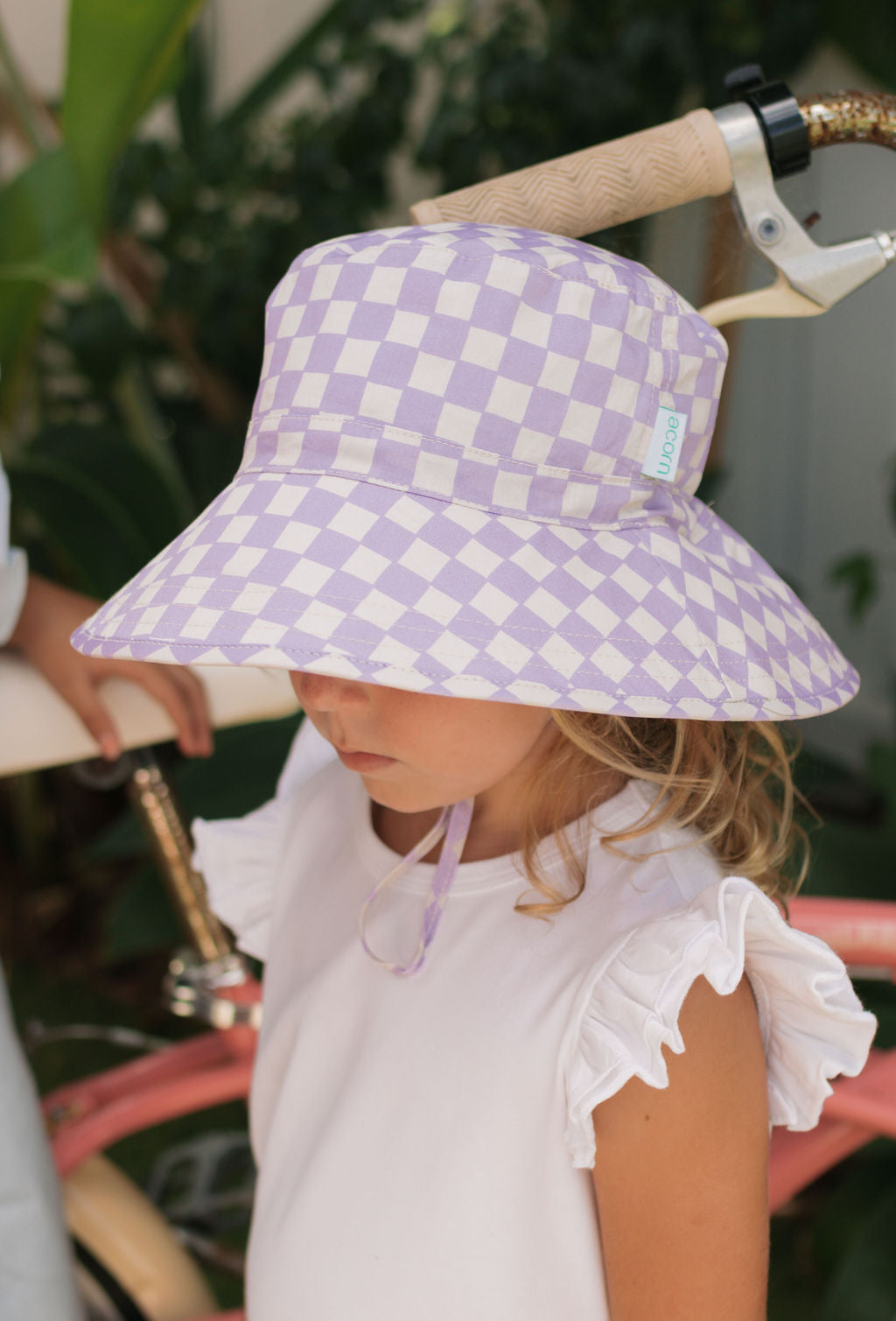A young child wearing the ACORN KIDS Acorn Kids - Lavender Checks Broad Brim Bucket Hat with UPF50+ sun protection and a white ruffled-sleeve top stands by a bicycle, the hat’s wide brim shading their face against lush greenery.