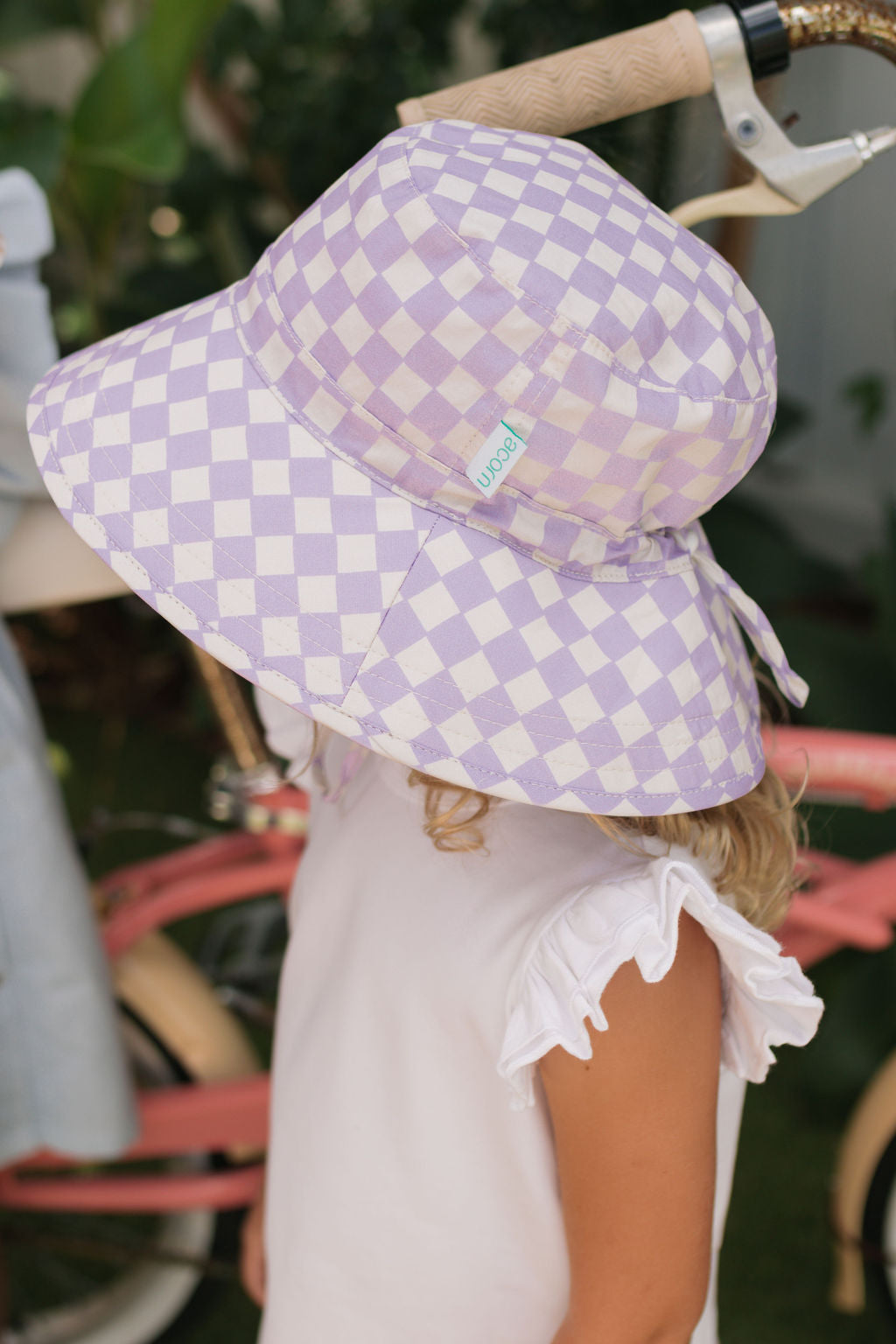 A young child in the ACORN KIDS Acorn Kids - Lavender Checks Broad Brim Bucket Hat with UPF50+ sun protection and a white ruffled shirt stands by a pink bike, their face mostly hidden by the oversized hat.