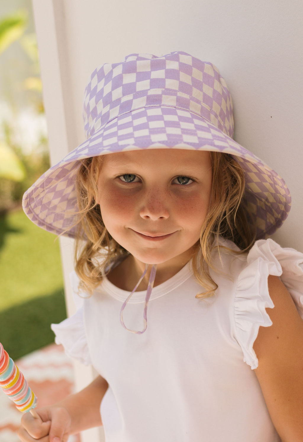 A young girl wears the ACORN KIDS Acorn Kids - Lavender Checks Broad Brim Bucket Hat and a white ruffled-sleeve top, smiling gently as she holds a colorful striped lollipop outdoors.