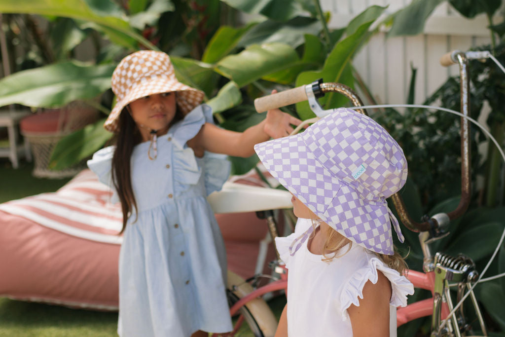 Two young girls stand outdoors near a bicycle. One wears a blue dress with a brown bucket hat, while the other sports a white dress with the ACORN KIDS Acorn Kids - Lavender Checks Broad Brim Bucket Hat (UPF50+). Lush plants and a pink lounge chair are behind them.