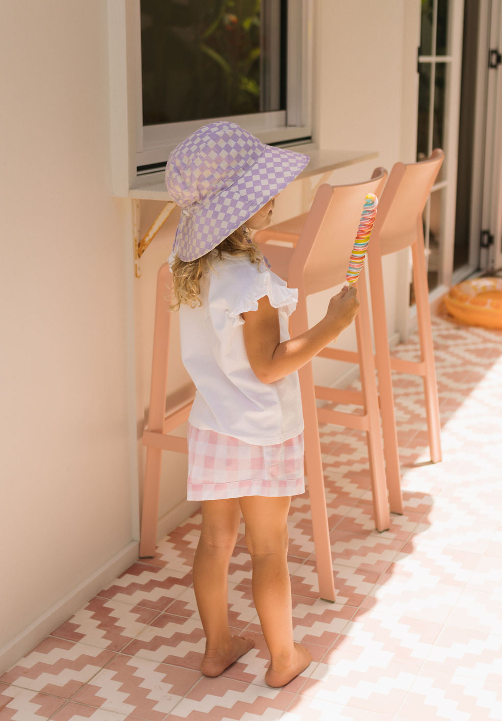 A young child stands barefoot on patterned tiles, holding a colorful ice cream while wearing the ACORN KIDS Acorn Kids - Lavender Checks Broad Brim Bucket Hat with UPF50+ sun protection near pink chairs and a window.