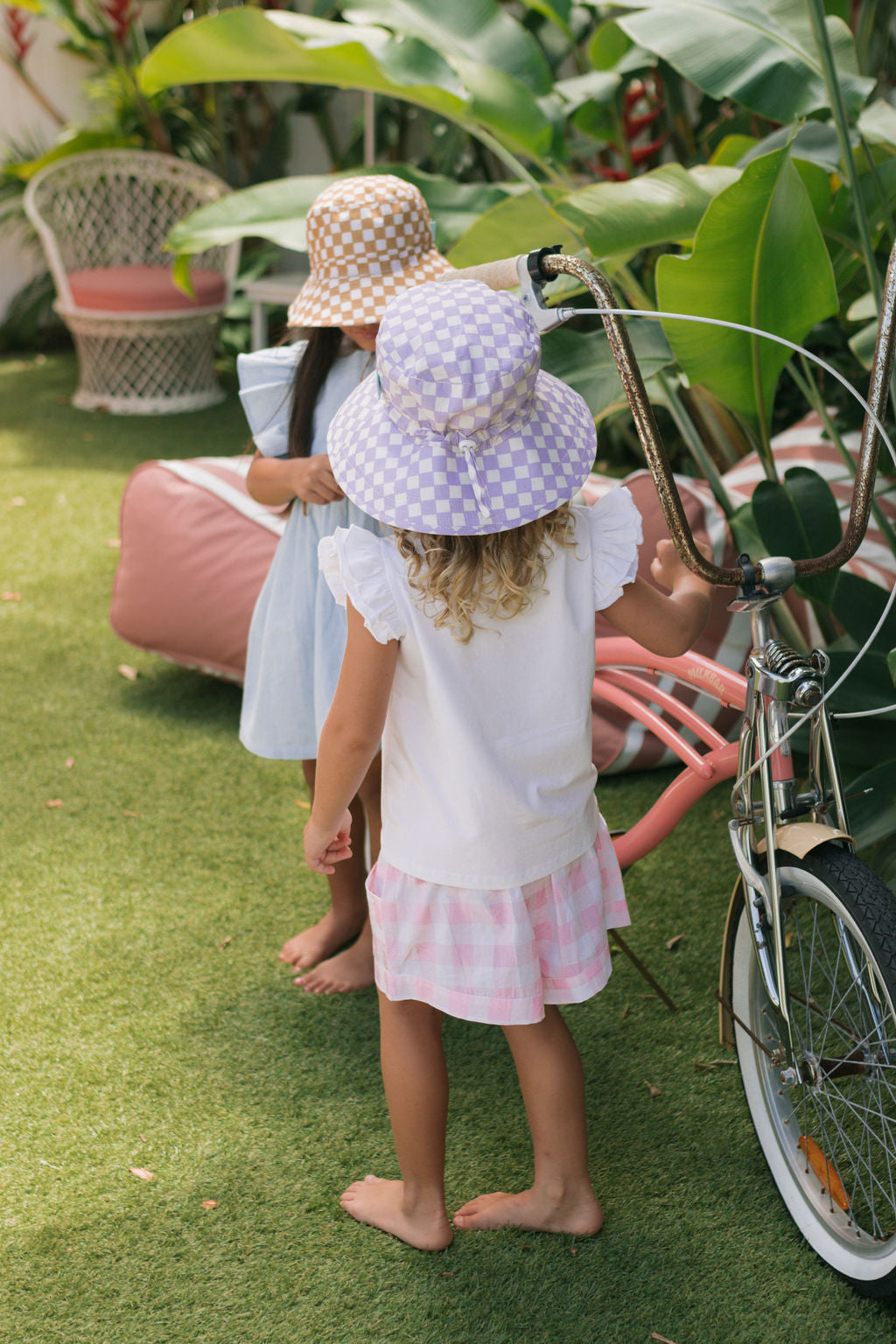Two young children wear ACORN KIDS' Lavender Checks Broad Brim Bucket Hats and light clothes as they stand barefoot on grass near a pink bench and bicycle, surrounded by lush green plants in a garden.