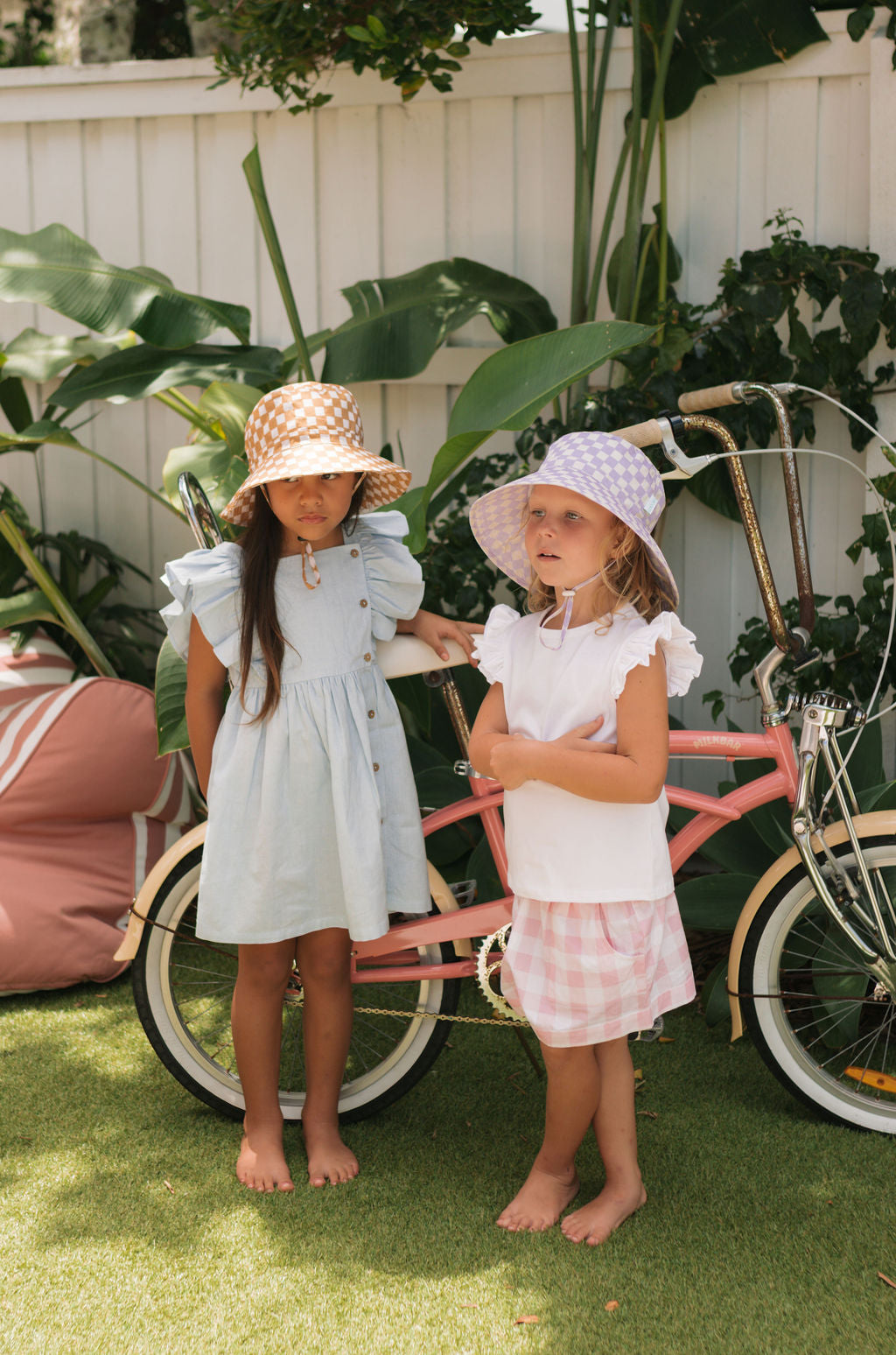 Two young girls barefoot on the grass stand by a pink bike, with green plants and a white fence behind. One wears a lavender checkered dress and the Acorn Kids - Lavender Checks Broad Brim Bucket Hat by ACORN KIDS for UPF50+ sun protection.