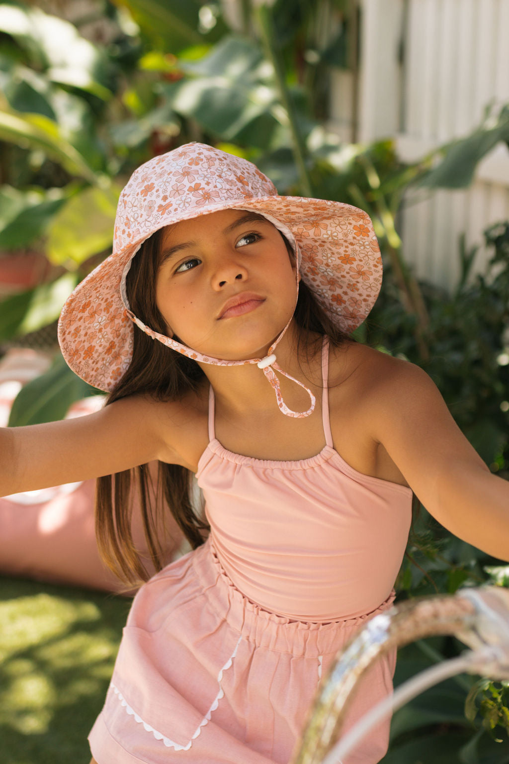 A young girl wears the ACORN KIDS Acorn Kids - Lulu Wide Brim Sunhat with a vintage floral print and a light pink dress, standing outdoors in a garden with lush green plants in the background.