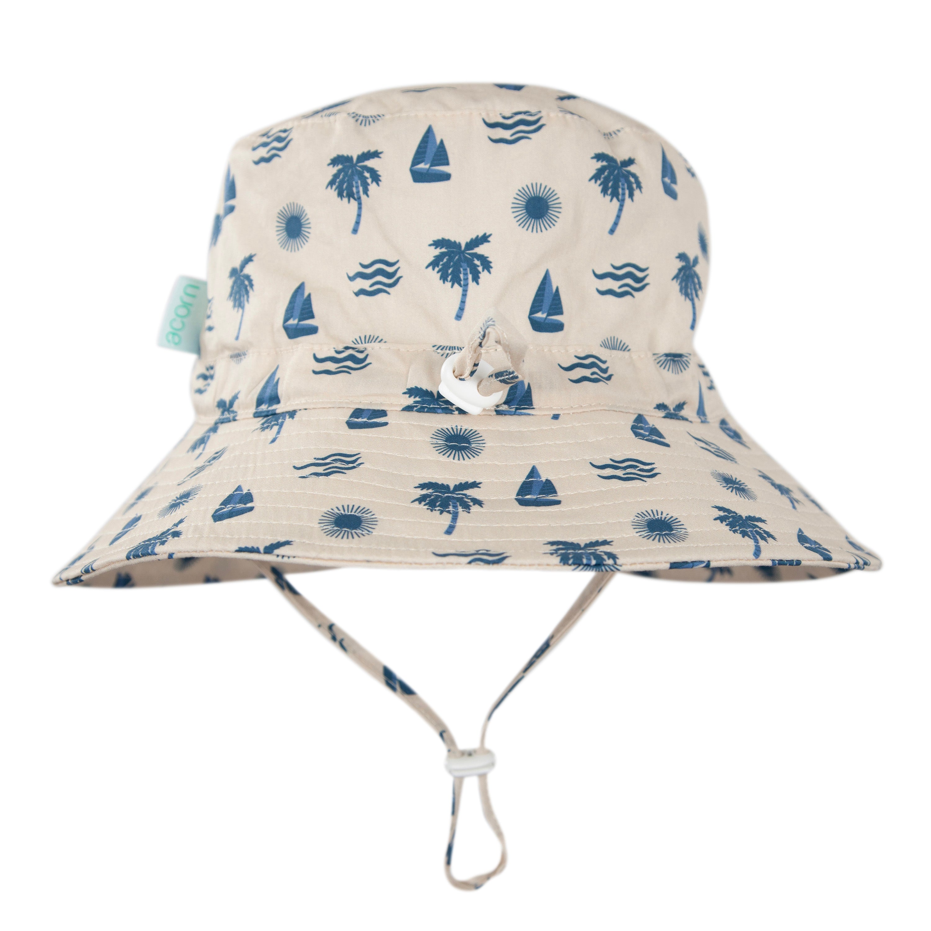 The Acorn Kids - Island Breeze Wide Brim Bucket Hat by ACORN KIDS is a beige cotton hat with blue palm tree, sailboat, wave, and sun prints. It offers UPF50+ sun protection, an adjustable chin strap, and a green fabric side tag.