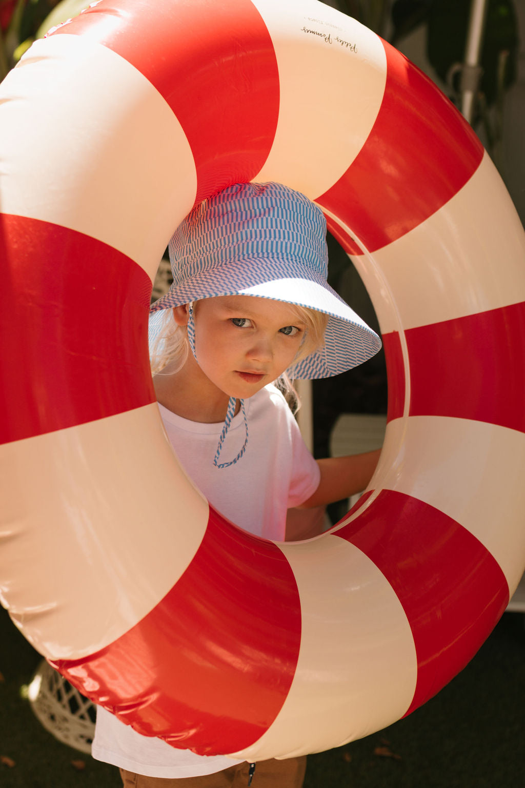 A young child wearing the Acorn Kids - Mykonos Wide Brim Bucket Hat by ACORN KIDS looks through a large red and white inflatable pool ring, sunlight casting warm tones on their face.
