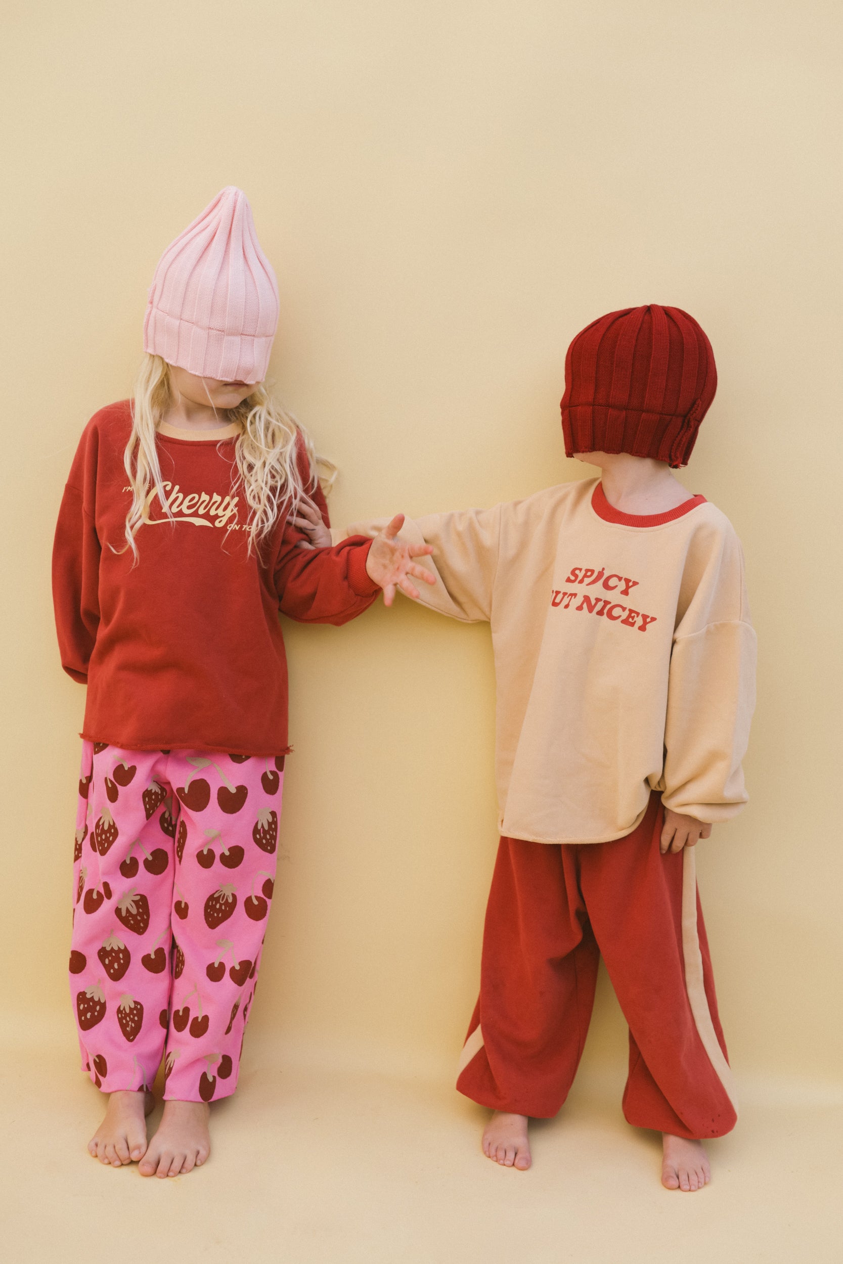 Two children stand barefoot against a beige background in colorful clothes and oversized knit hats. One wears pink strawberry pants and a red T-shirt jumper; the other sports best-selling red pants and SMALL SWIM CLUB's Spicy But Nicey Pullover.