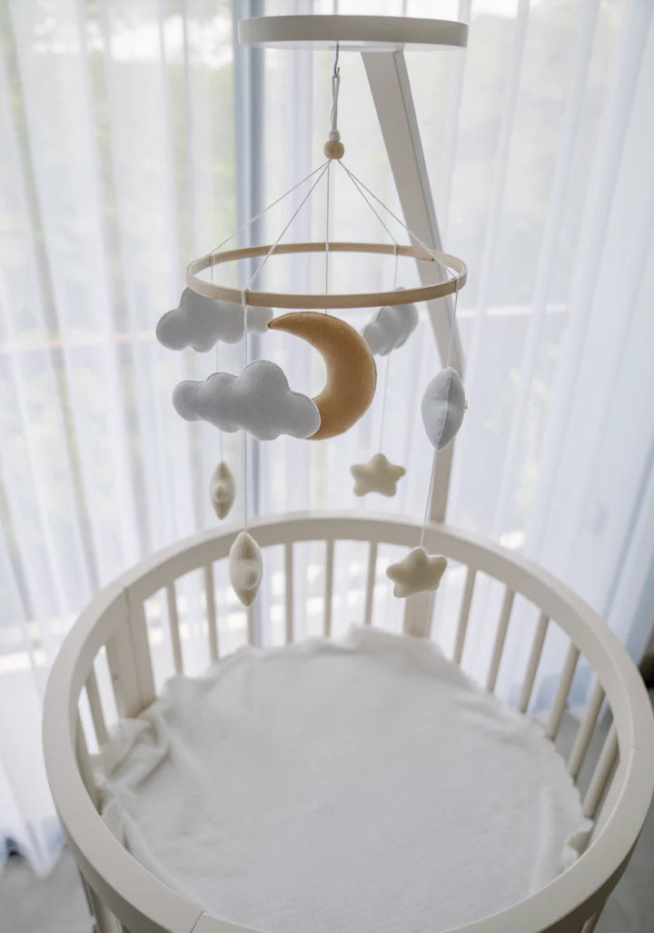 A round white baby crib with a soft mattress is topped by the ROOMI & ME - Baby Mobile Starry Night, featuring handmade clouds, stars, and a crescent moon for gentle visual stimulation in front of sheer curtains and a bright window.