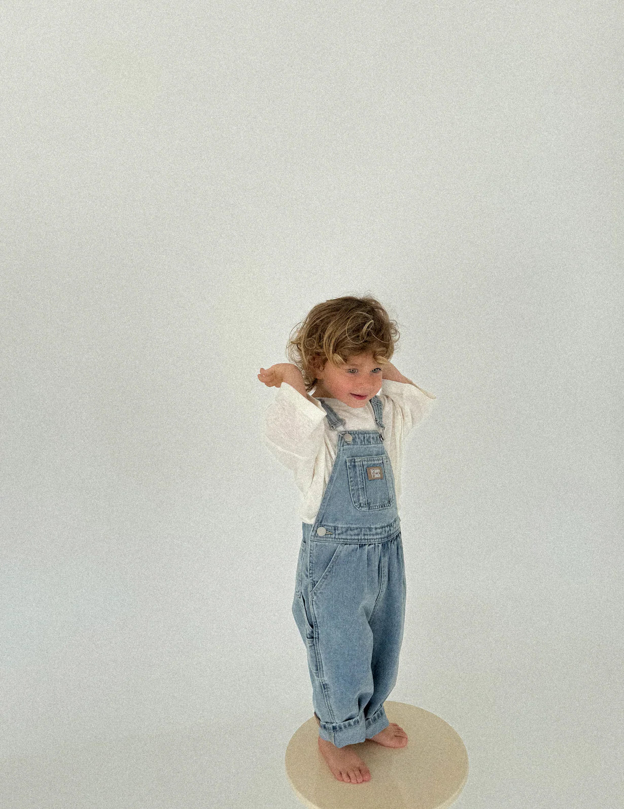 A young child with curly hair wears TWIN COLLECTIVE’s Twin Collective - Carpenter Overall and a white shirt, standing barefoot on a stool against a plain light background, hands behind their head.