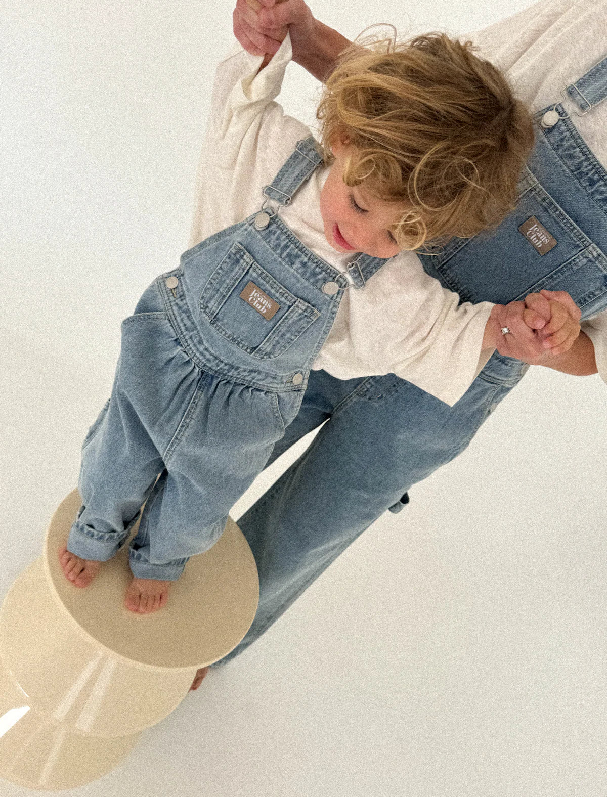 A young child in TWIN COLLECTIVE’s Carpenter Overall stands barefoot on a beige stool, holding hands with an adult in matching overalls, both set against a plain light background.