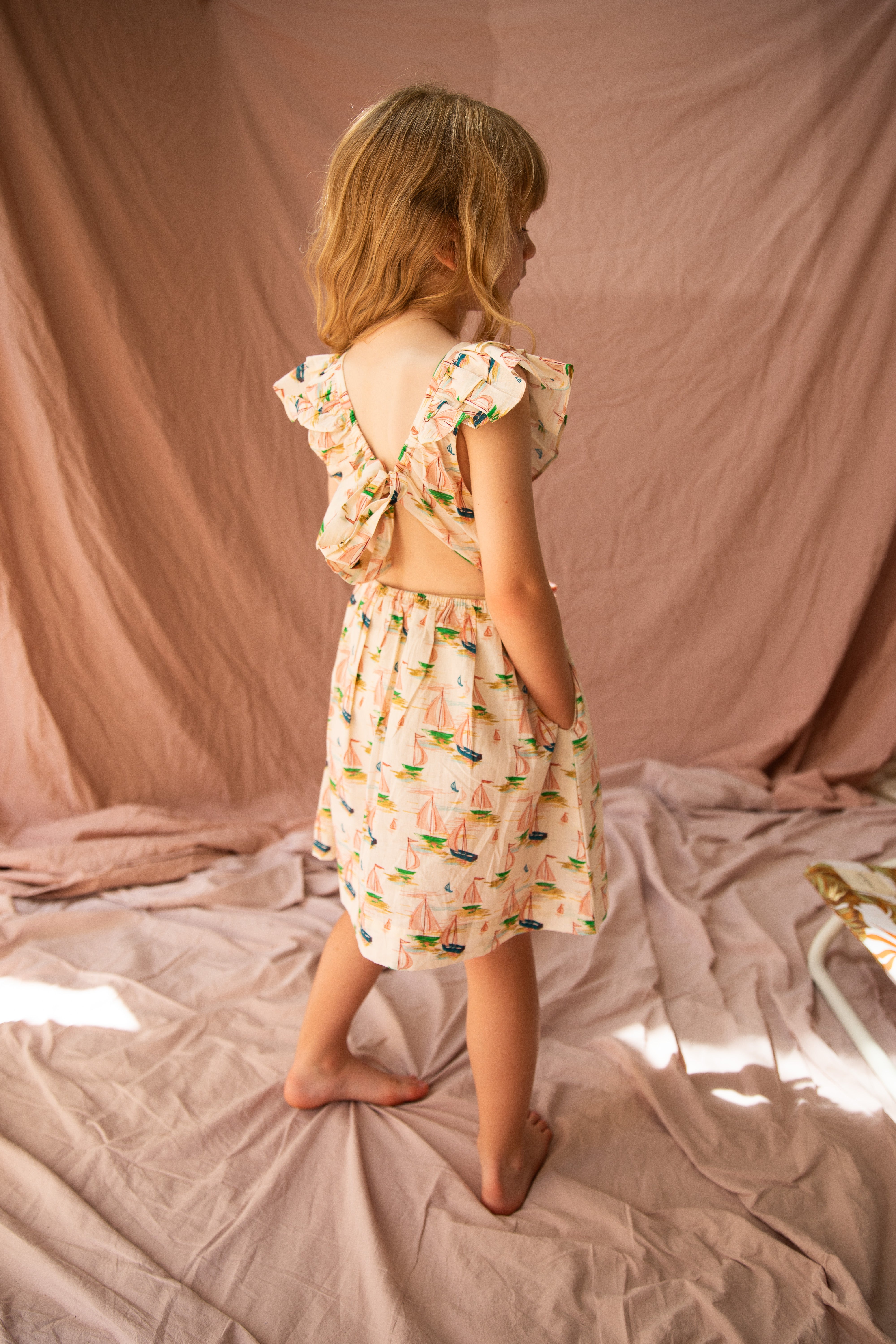 A young girl with blonde hair stands barefoot on a pink fabric backdrop, illuminated by warm lighting, wearing the sleeveless, open-back Seychelles Dress Regatta by BELLA + LACE—an ideal summer outfit featuring a vibrant print.