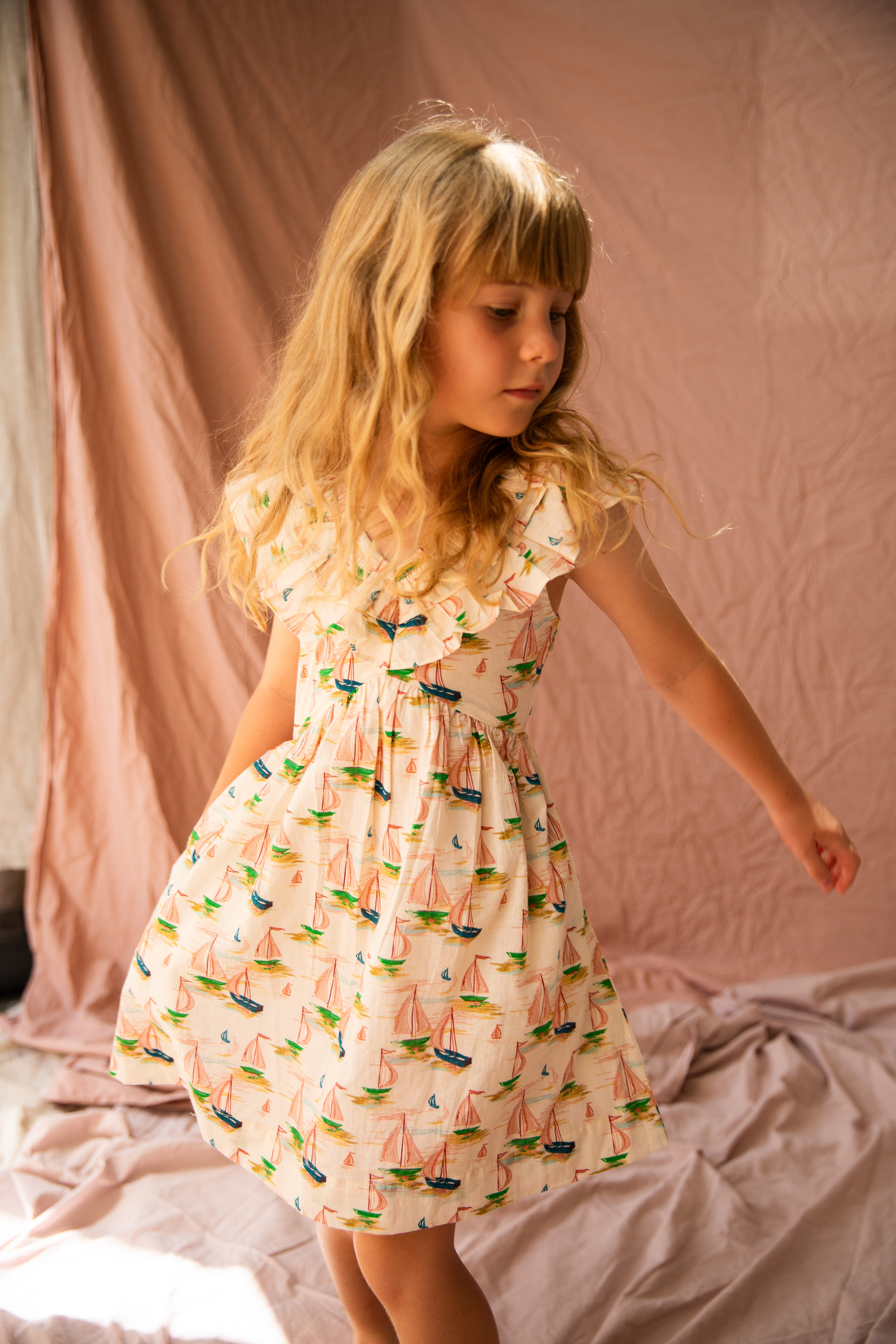 A young girl with long blonde hair twirls in the BELLA + LACE Seychelles Dress Regatta, which is adorned with a colorful print of sailboats. The background consists of soft, neutral-colored cotton voile drapes, and light casts gentle shadows on the scene.
